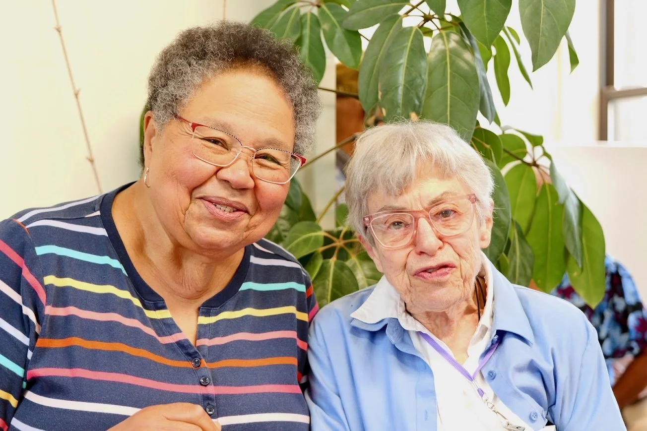 two older women are sitting in front of a plant smiling at the camera