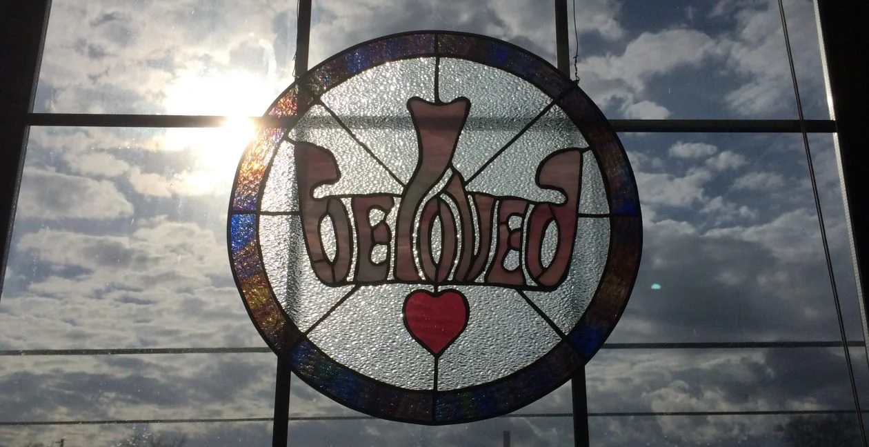 a stained glass piece is handing in the window with the sun shining behind it and reads Beloved with a heart under it