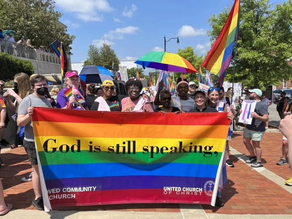 a group of people is behind a large rainbow sign that reads "God is still speaking"