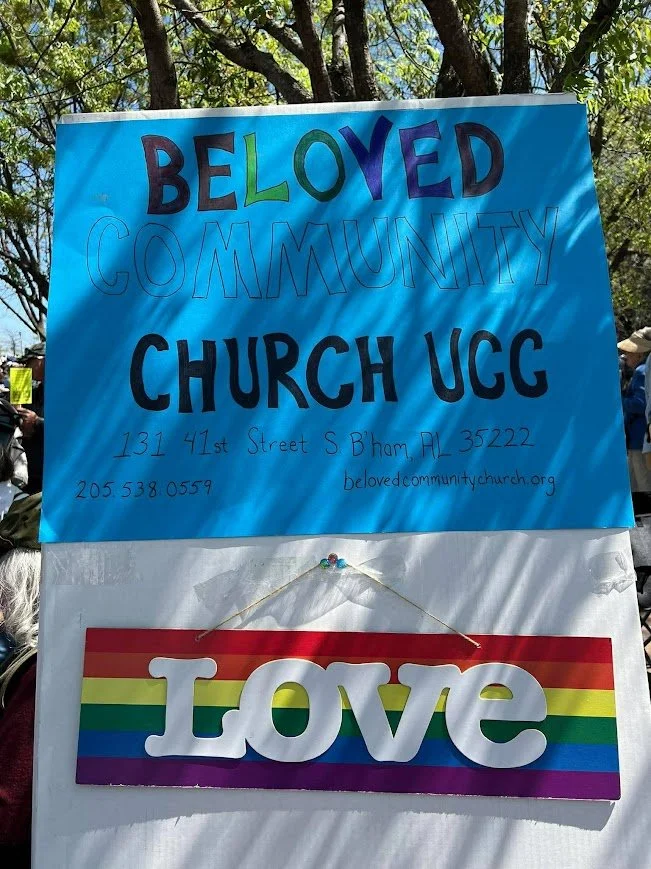 a sign that is outdoors is blue and reads Beloved Church UCC and there is a rainbow flag that says LOVE under it