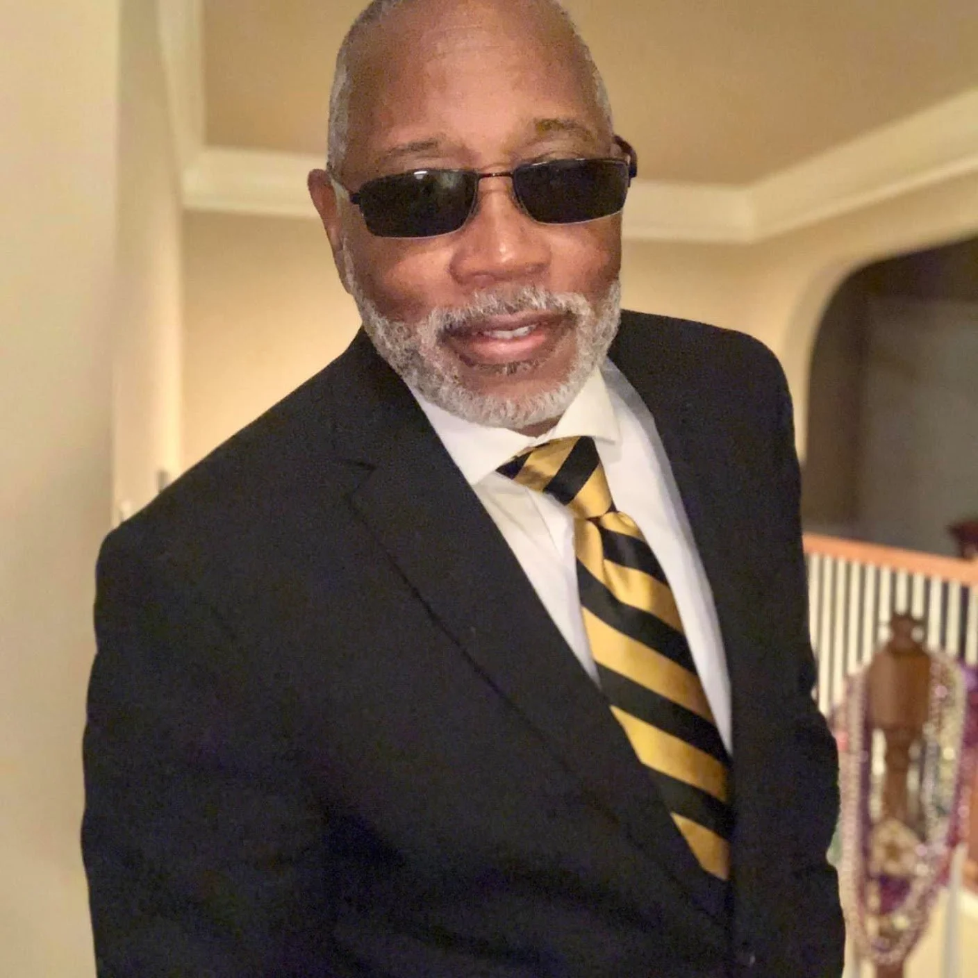 a Black man with sunglasses and wearing a suit looks at the camera smiling
