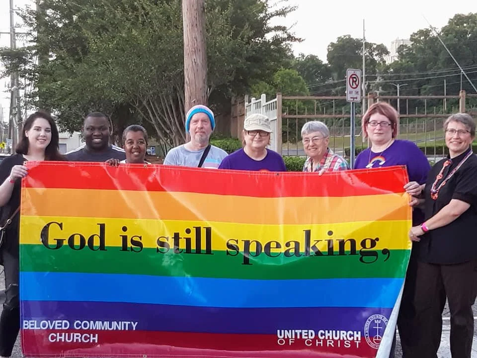 a group of people stands behind a large rainbow banner that reads God is still speaking,