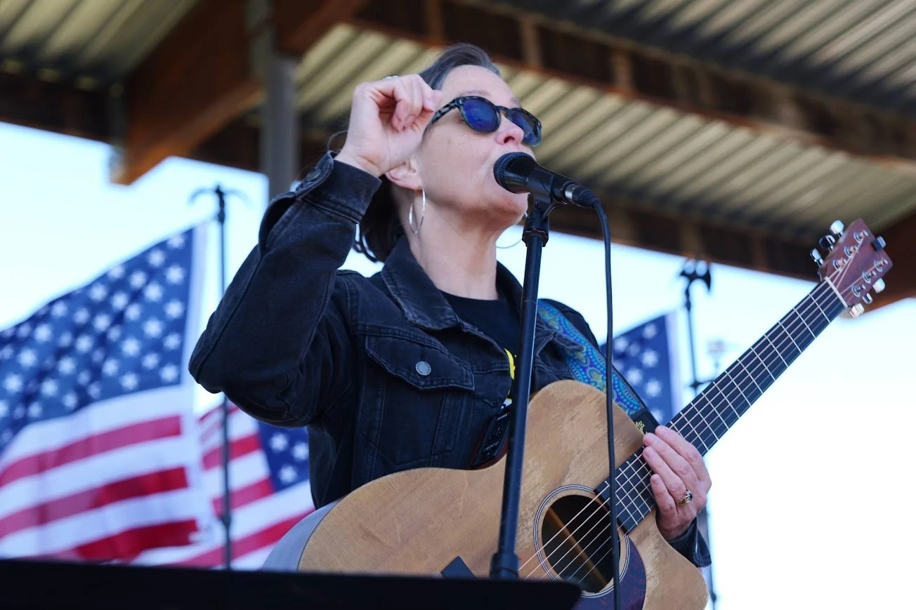 a person is wearing sunglasses, singing at a mic holding a guitar in front of an American flag