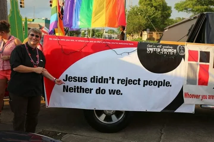 a person is standing next to a sign pointing at it, the sign reads Jesus didn't reject people. Neither do we.