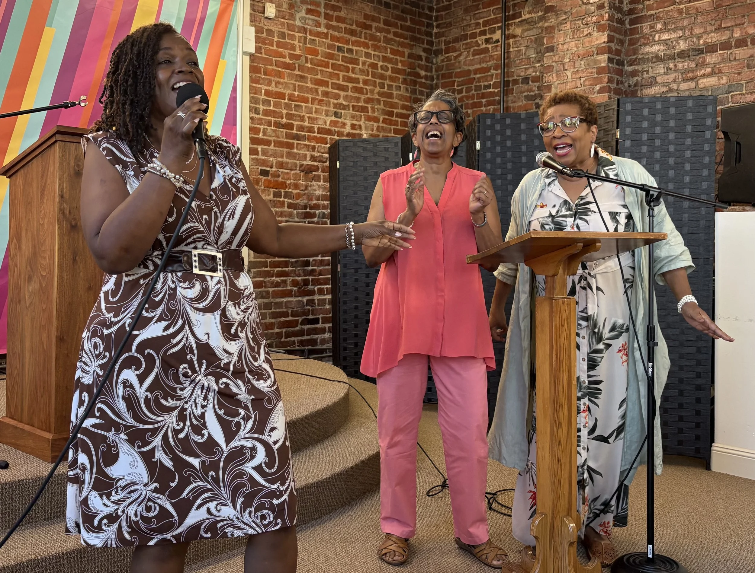 Three Black women are singing with microphones and a pulpit behind them