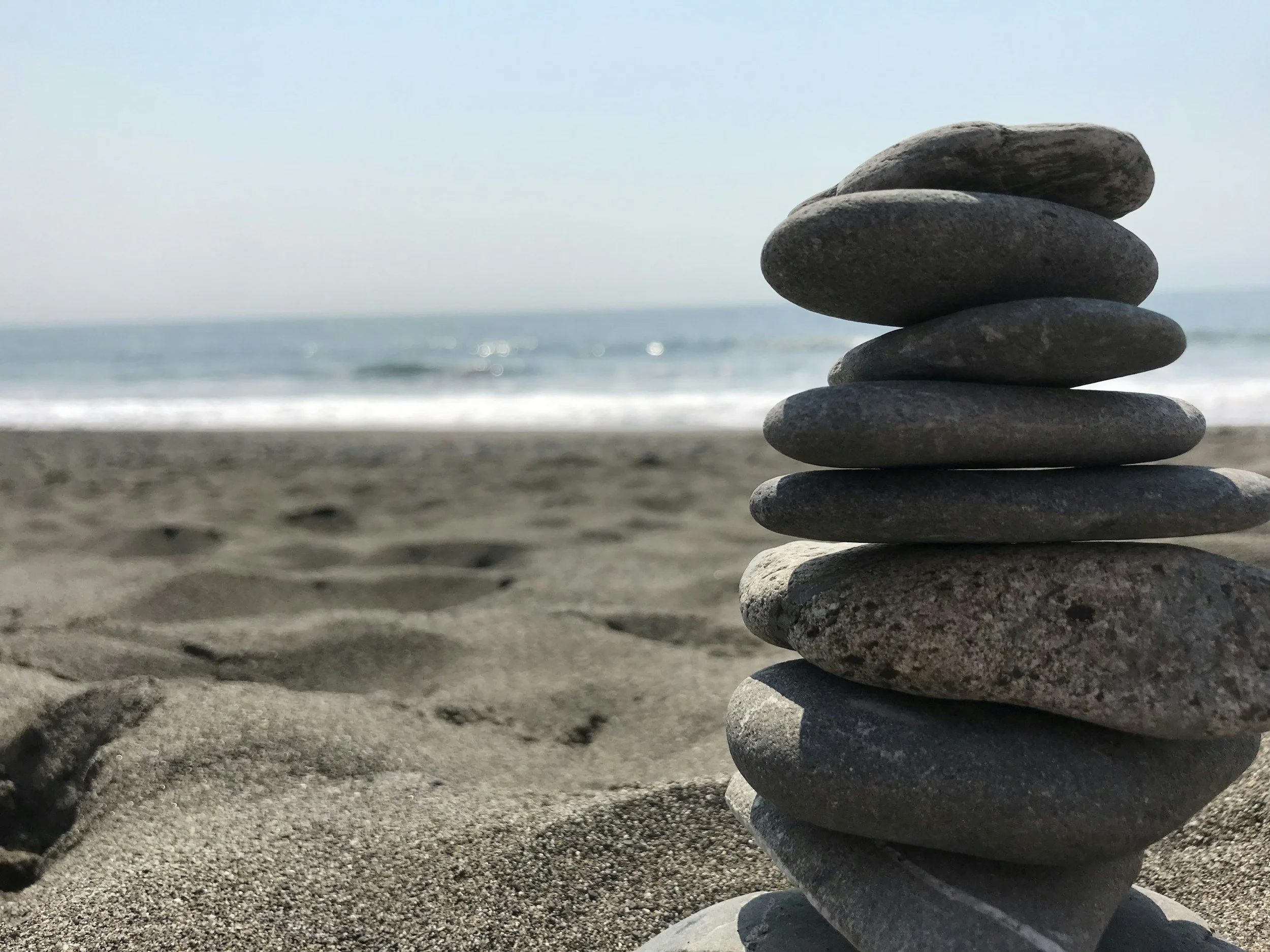 Stack of seven smooth, flat stones balanced on a sandy beach with ocean waves and a clear sky in the background.