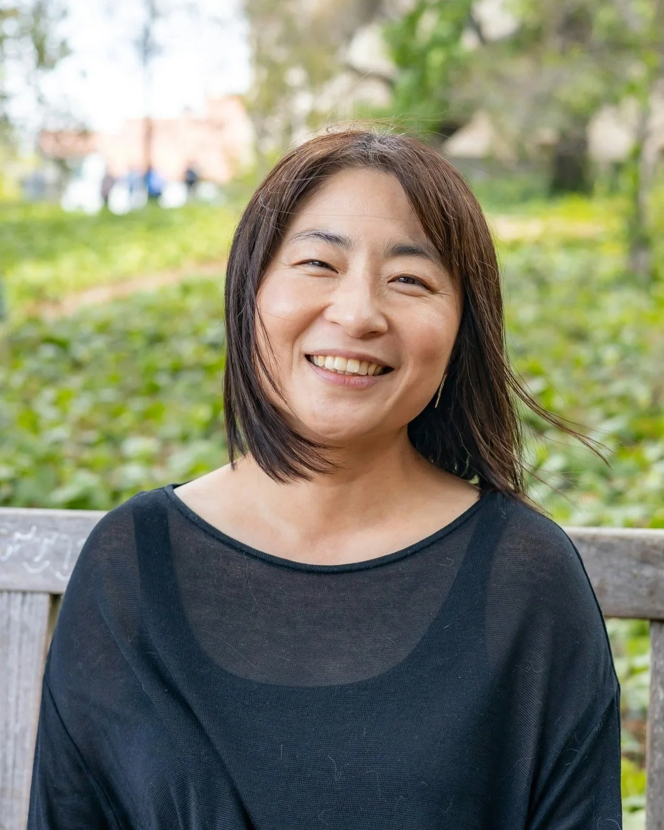 A woman smiling and sitting on a park bench with greenery and trees in the background.