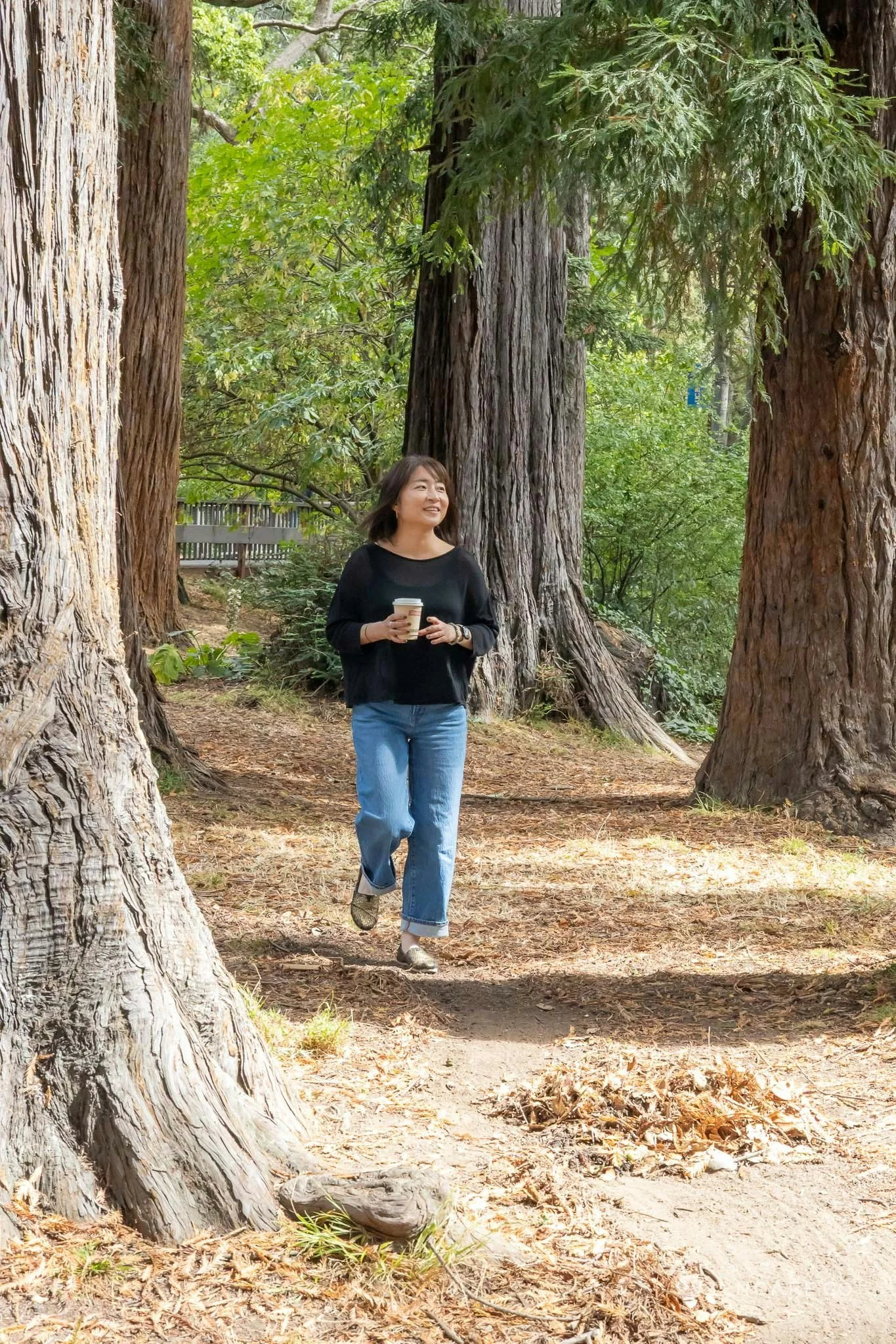 A woman walking along a forest trail, holding a coffee cup and smiling, surrounded by tall trees and green foliage.