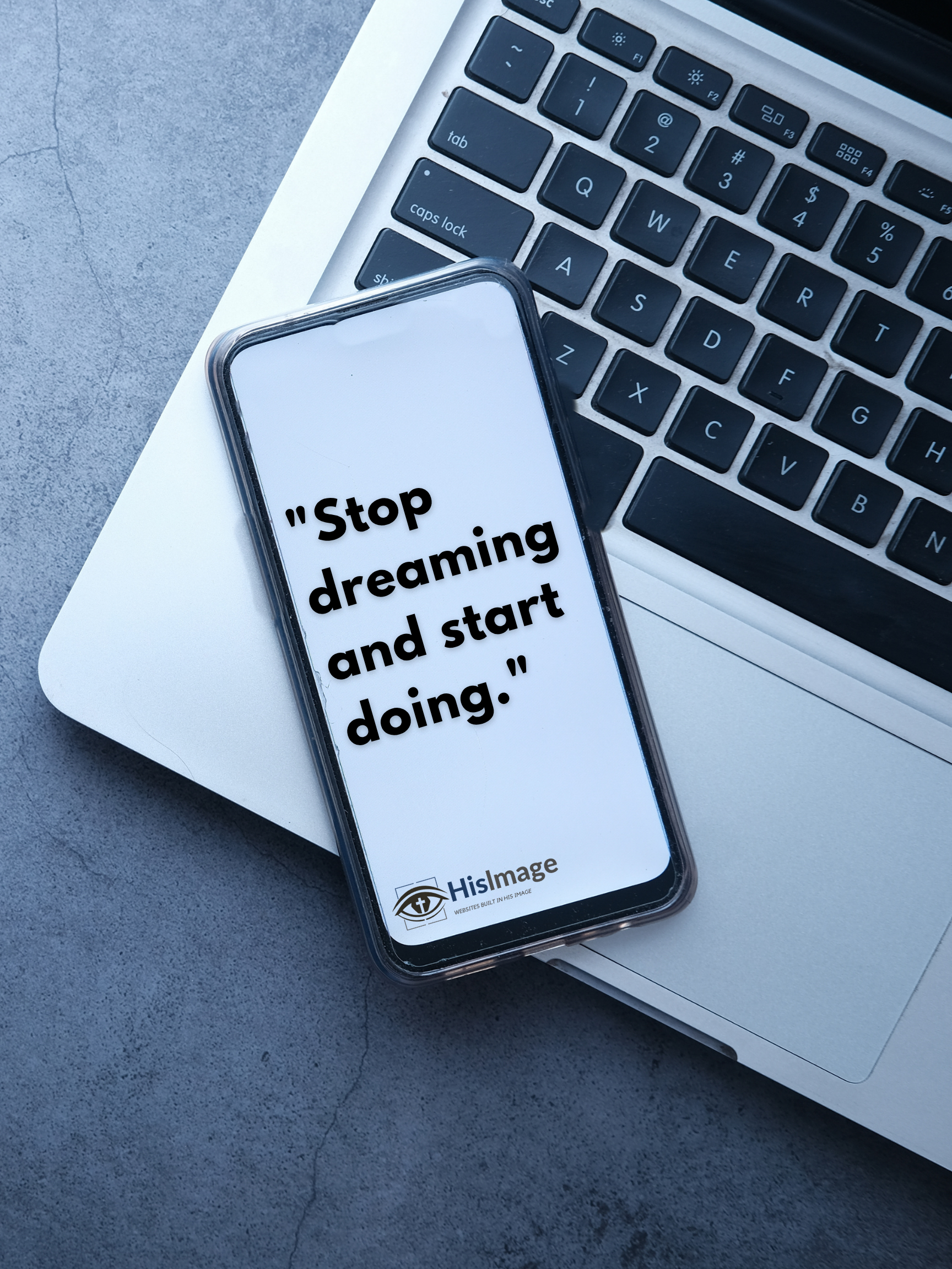 Close-up of a laptop keyboard with a smartphone placed on top of it. The screen of the smartphone displays a quote: 'Stop dreaming and start doing.' The phone has a clear case and is positioned on a white sheet of paper, with a gray textured surface underneath.