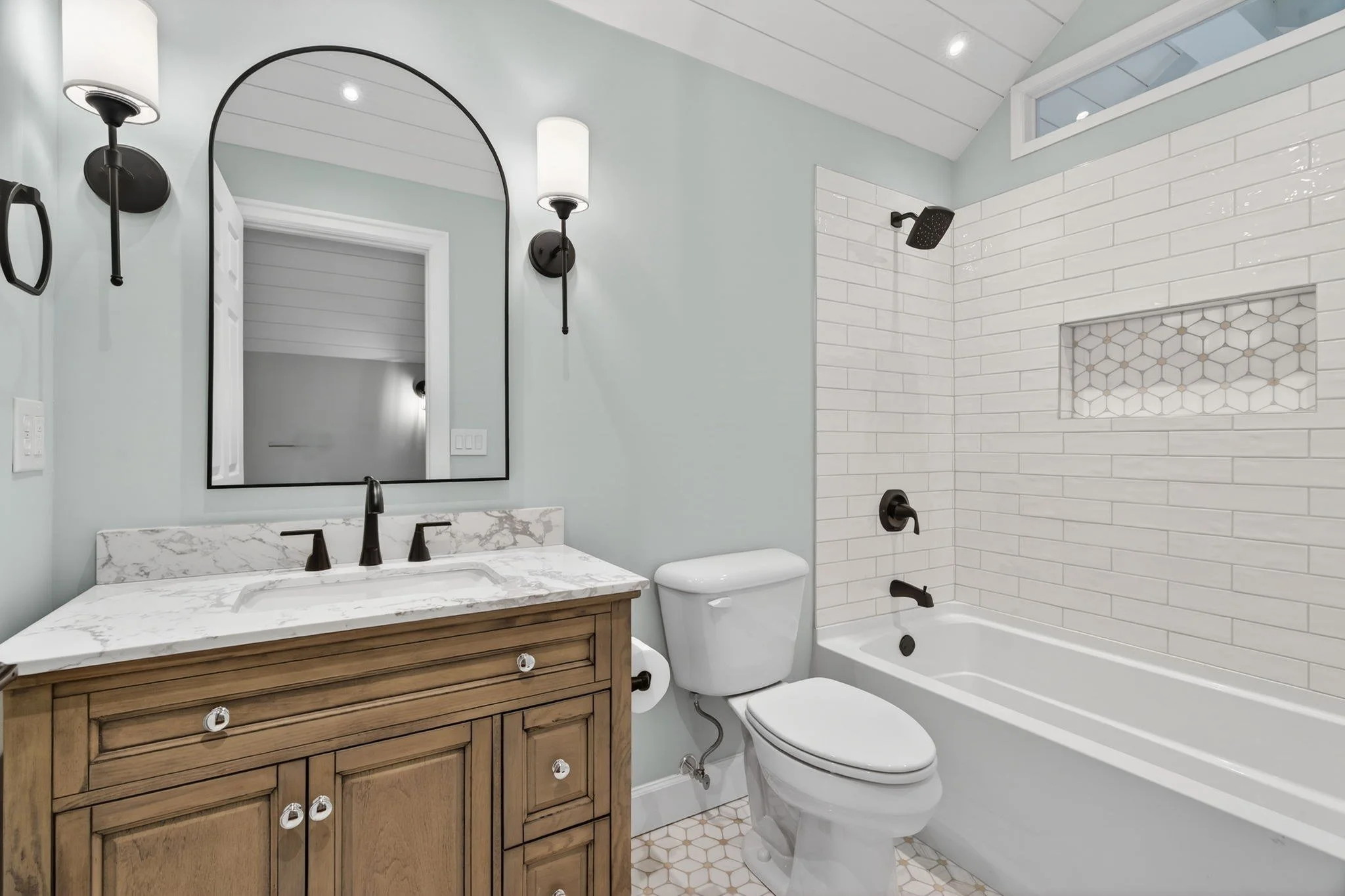 Sage green bath with a wood vanity, arched mirror, and subway tile tub surround