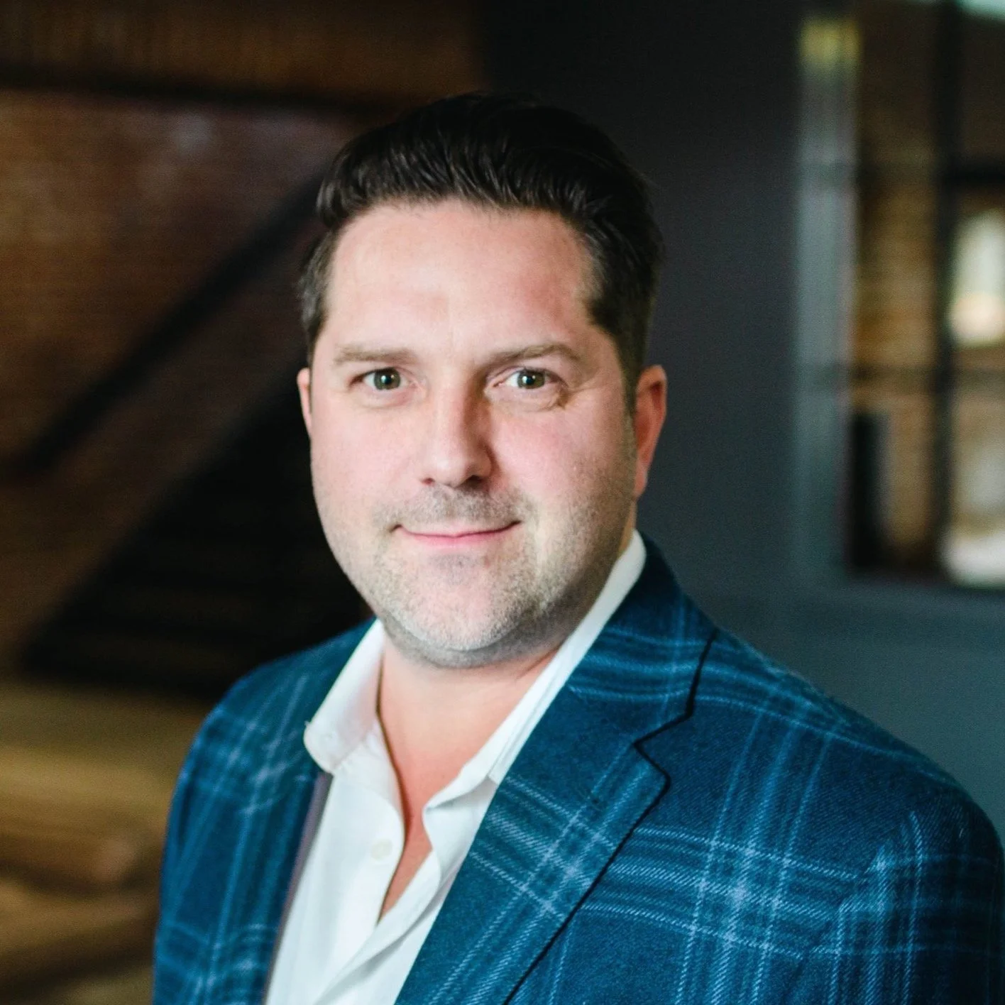 Professional headshot of a man with dark hair, wearing a blue checkered blazer and a white shirt, standing in front of a wooden background.