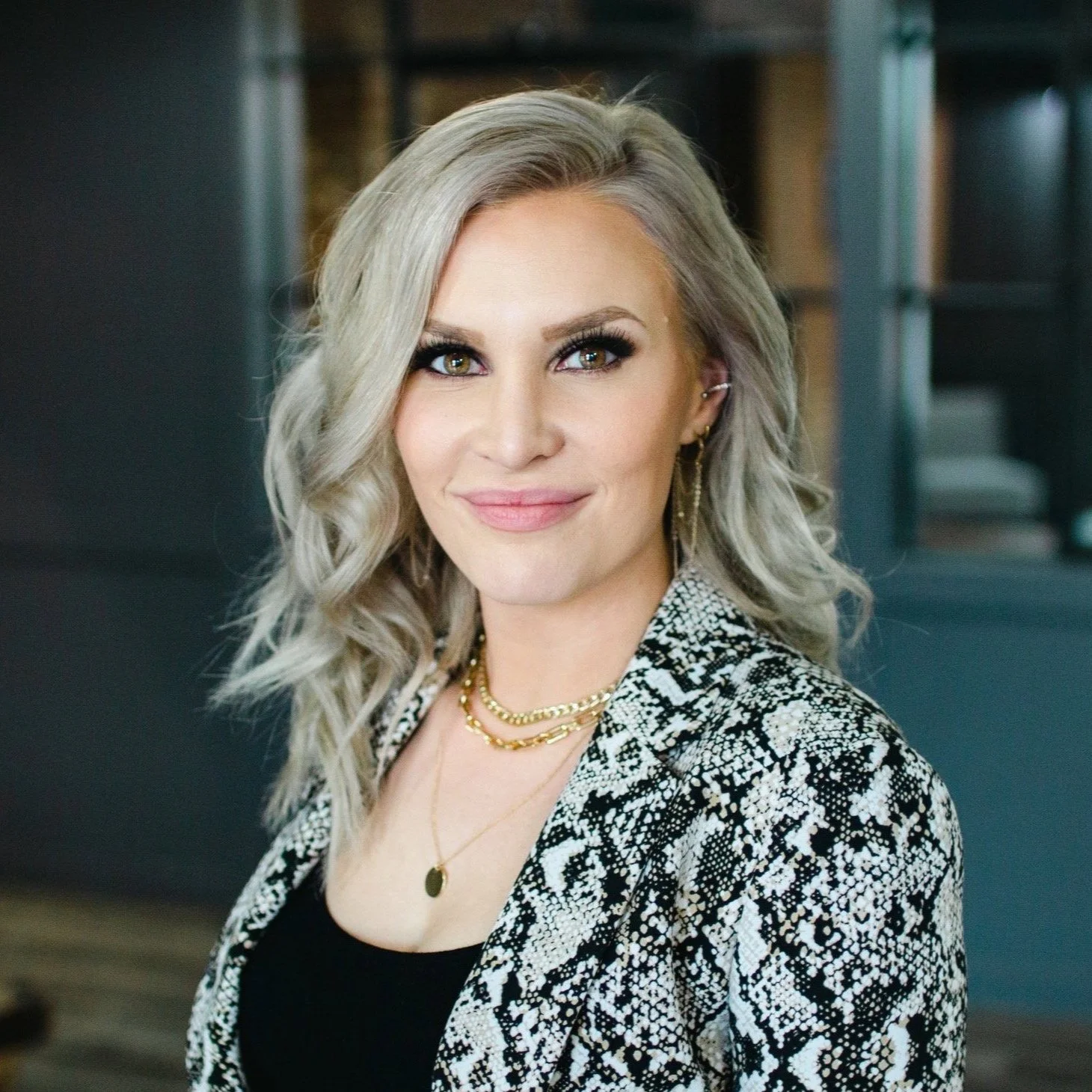 A woman with shoulder-length blonde hair, wearing a black top and a black and white patterned blazer, smiling in an indoor setting with modern furniture and a dark background.