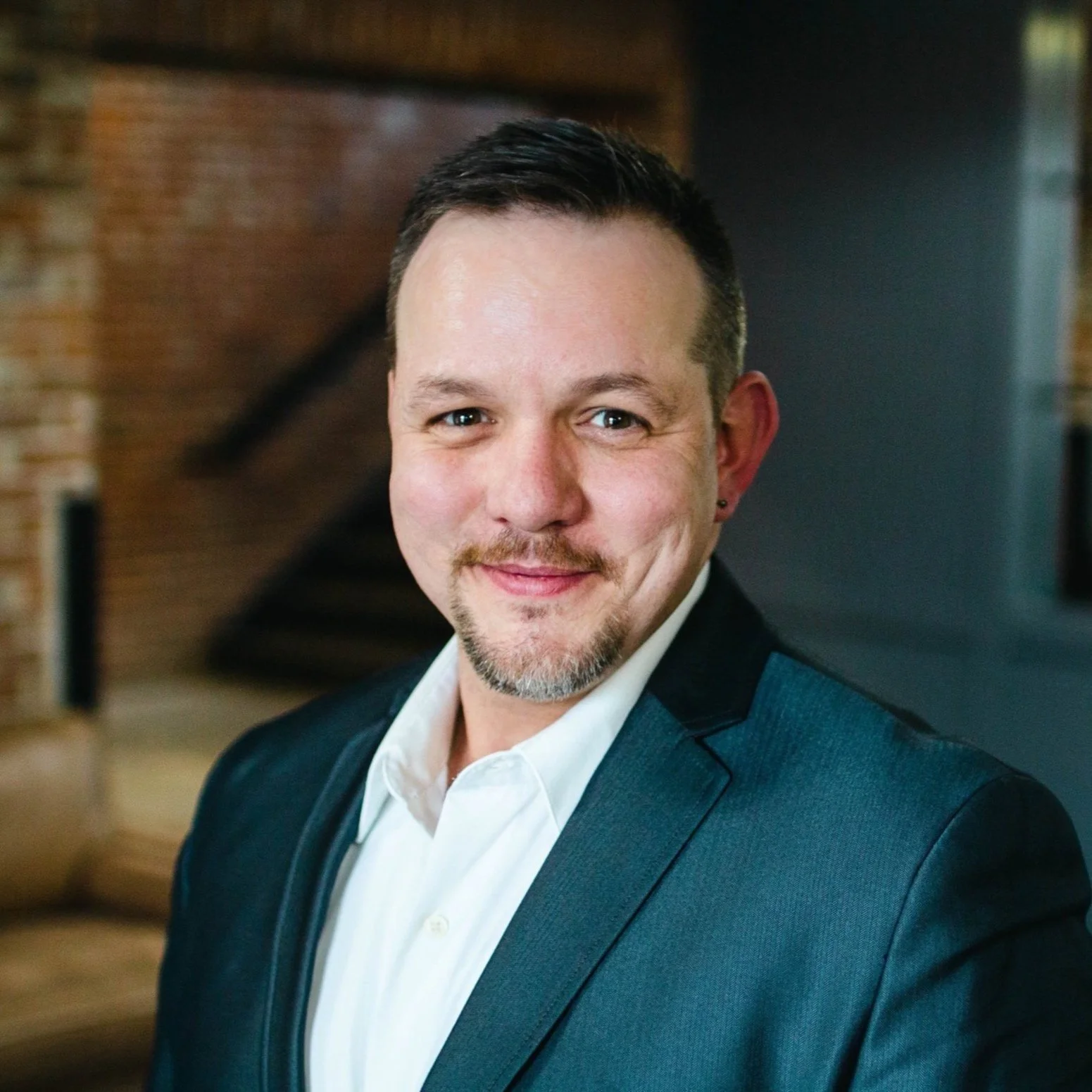 Headshot of a smiling man with short dark hair, a goatee, and wearing a dark suit jacket over a white shirt, in an indoor setting with brick walls and staircase in the background.
