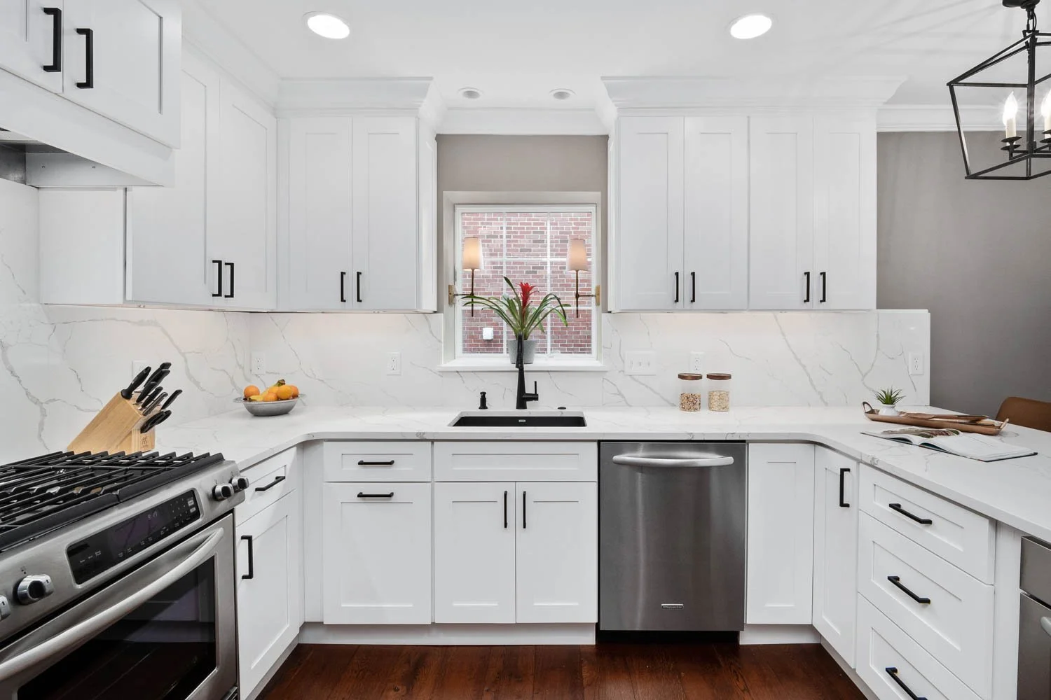 Modern white kitchen with black hardware, marble countertops, stainless steel dishwasher, and stove, with a window above the sink, decorative jars, and small plants.