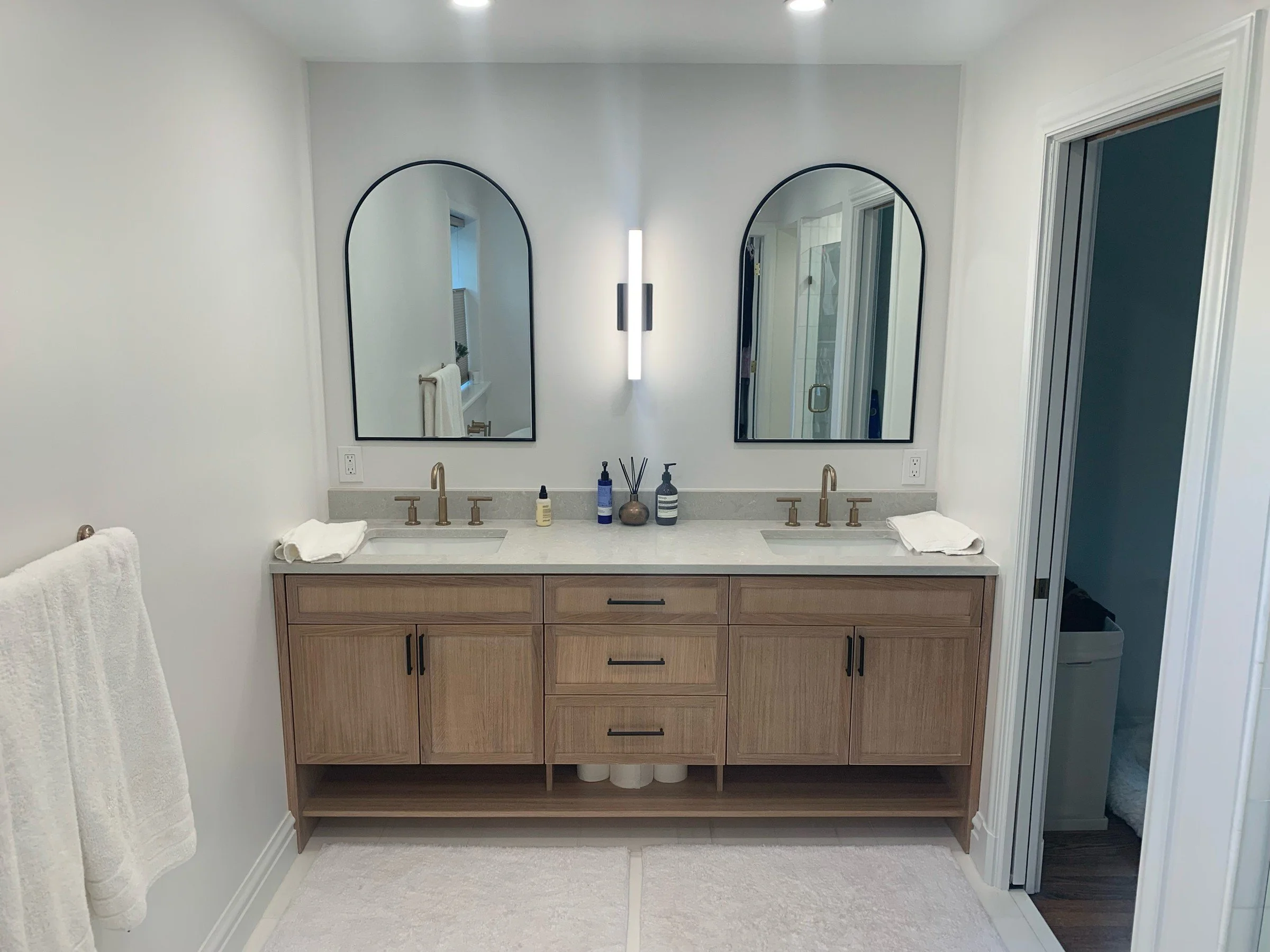 Double bathroom vanity with white oak cabinets, a quartz top, brass faucets, and arched black mirrors