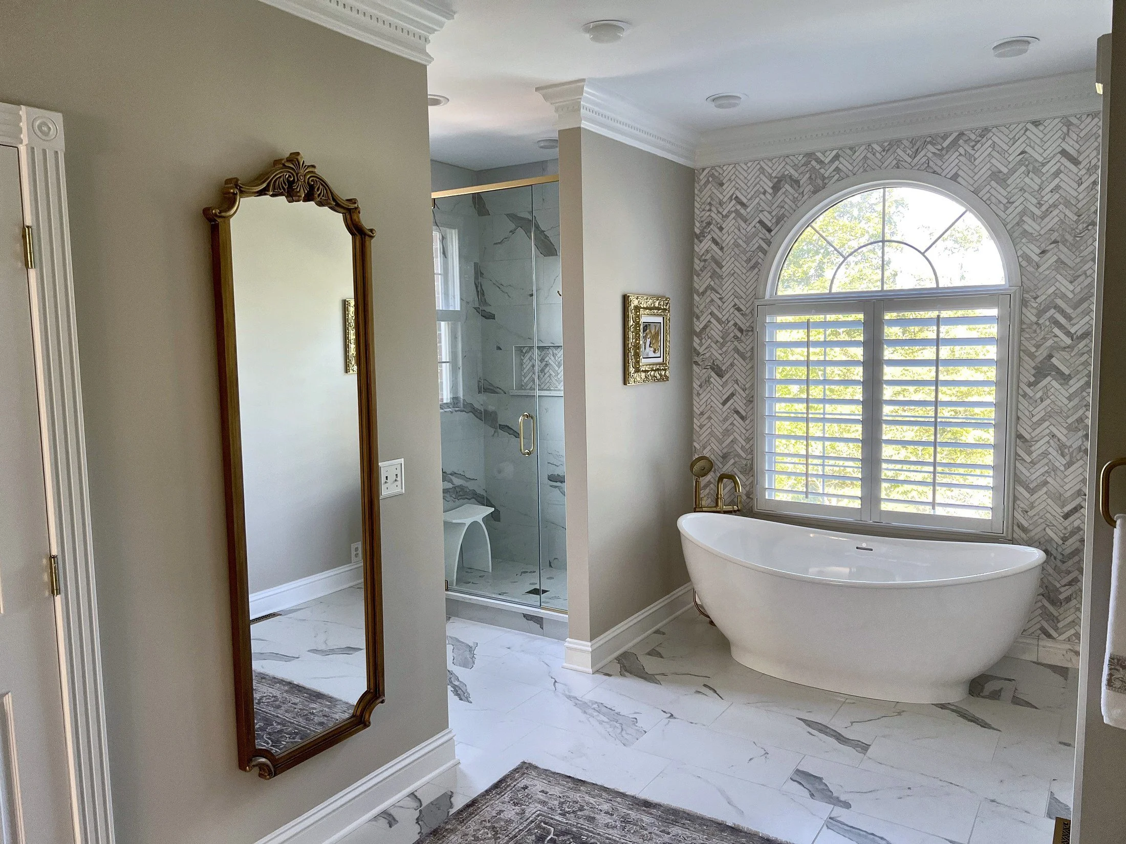 Freestanding tub, frameless glass shower, and an antique mirror in a primary bath with a herringbone tile wall