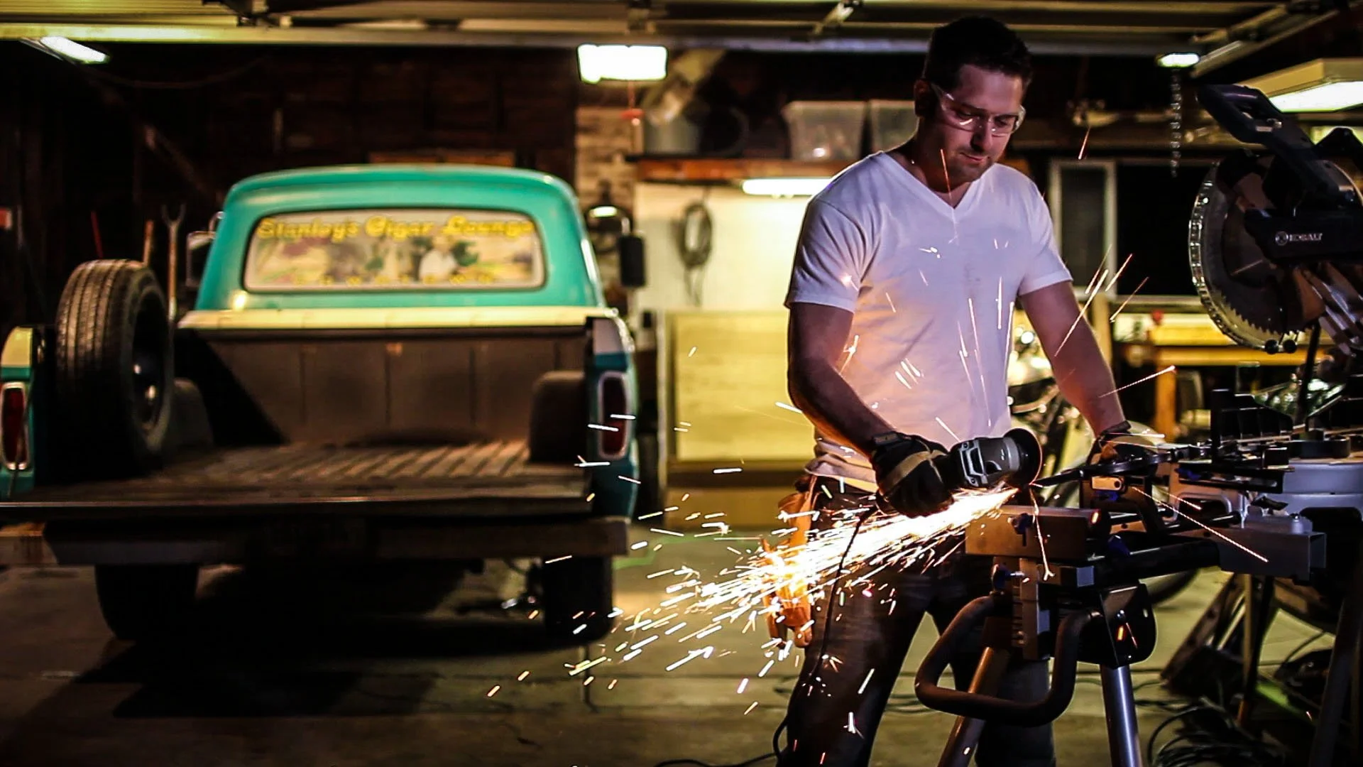 A man in a white t-shirt and safety glasses welding metal with sparks flying in a workshop. A vintage green pickup truck is parked in the background.