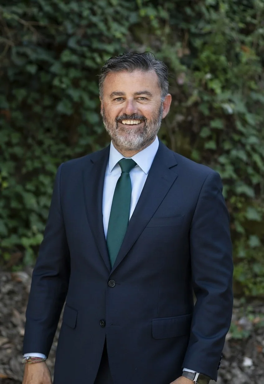 Hombre de negocios con barba y cabello gris, vestido con traje azul y corbata verde, de pie al aire libre con fondo de vegetación.