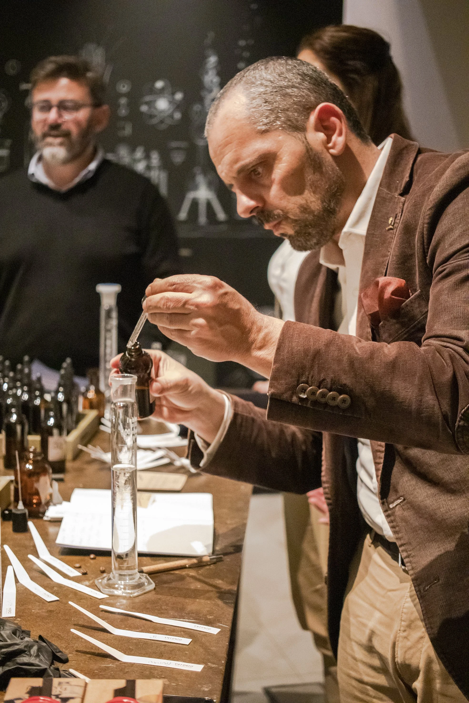 Hombre realizando un experimento con tinturas y frascos en una mesa de laboratorio o taller, con otras personas observando.