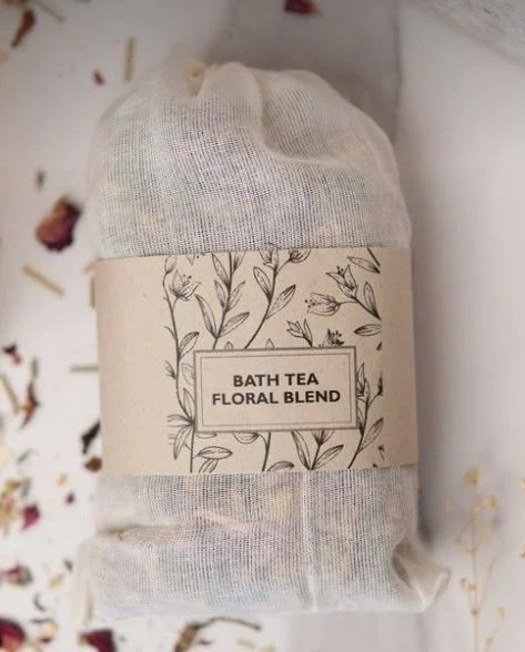 A rolled-up tea bag labeled 'Bath Tea Floral Blend' with a floral design on the packaging, placed on a surface surrounded by dried flower petals.