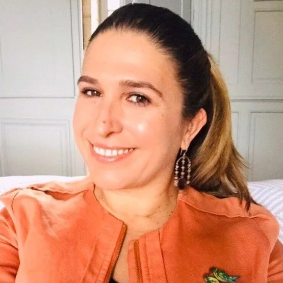 Mujer sonriendo con cabello recogido, lleva una chaqueta color durazno y pendientes grandes, y está en un ambiente interior.