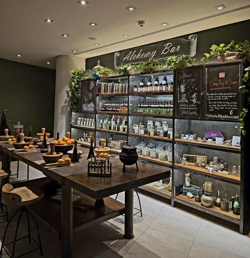 Barra de ingredientes y productos naturales en un espacio llamado 'Alchemy Bar', con estanterías llenas de frascos, plantas y pizarras con instrucciones y explicaciones en una cafetería o tienda especializada en productos de bienestar y spa.