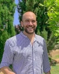 Hombre sonriendo con barba, camisa de rayas, al aire libre con árboles y cielo en el fondo.