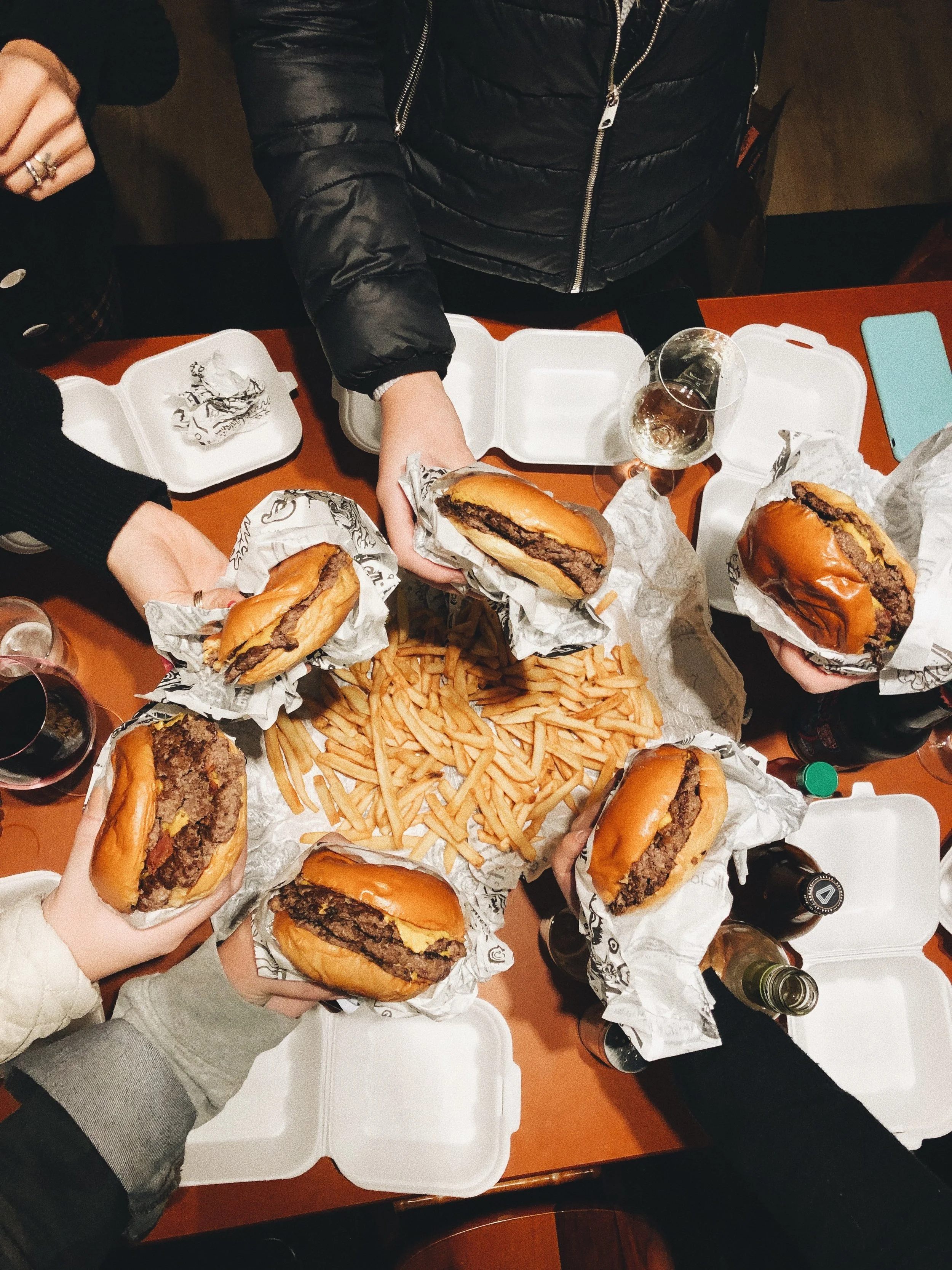 People at a table holding cheeseburgers wrapped in paper, with a large pile of French fries and bottles of soda and beer on the table.