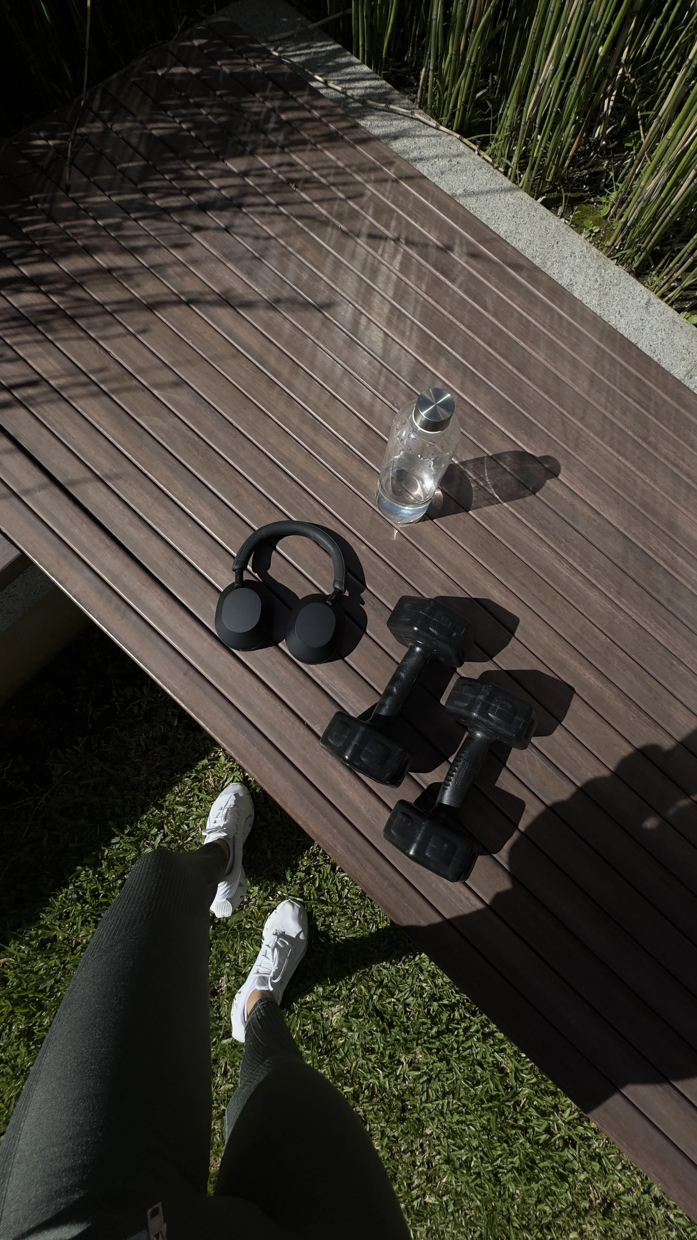 A person's legs in black leggings and white sneakers are visible, standing on grass beside a wooden bench. The bench has a water bottle, a pair of black headphones, and two black dumbbells placed on it. The scene is outdoors with sunlight and shadows.