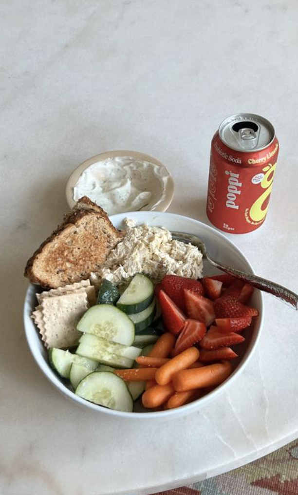A bowl of fresh vegetables including cucumber slices, strawberries, baby carrots, and sliced turkey, accompanied by a slice of bread with butter, a scoop of chicken salad, and a small dish of ranch dressing. A can of cherry vanilla soda is next to the bowl.