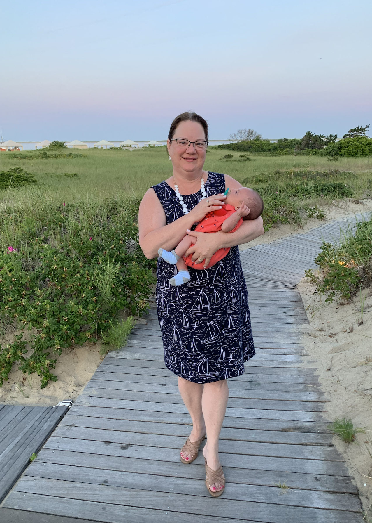 A woman with glasses and a necklace is walking on a wooden boardwalk on a beach while holding a baby in her arms. There are green bushes and a grassy area in the background with beach umbrellas visible in the distance under a sky with light pastel colors.