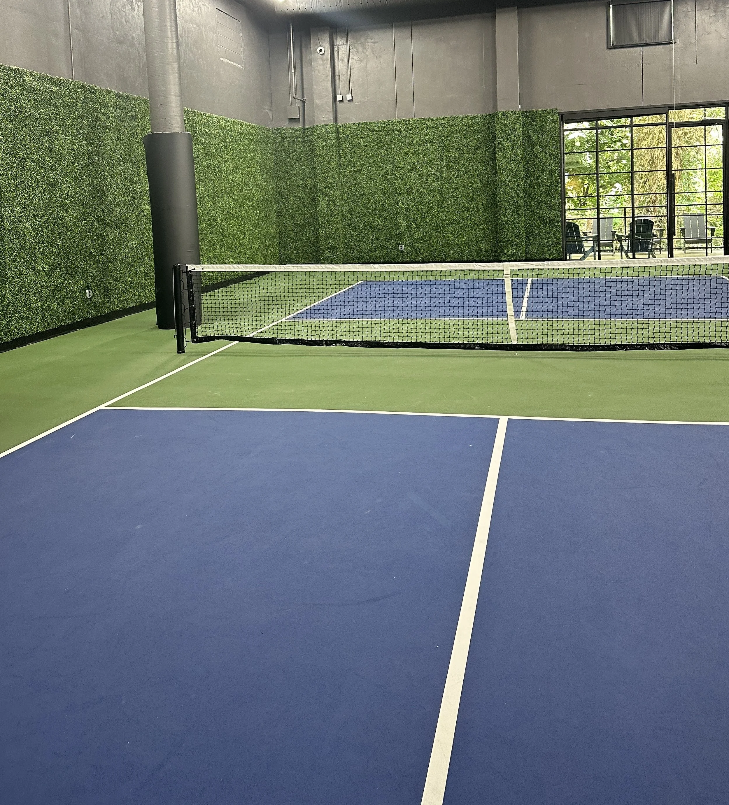Indoor tennis court with a blue and green surface, a black net, and a green artificial hedge wall. Large glass doors show outdoor seating and greenery.