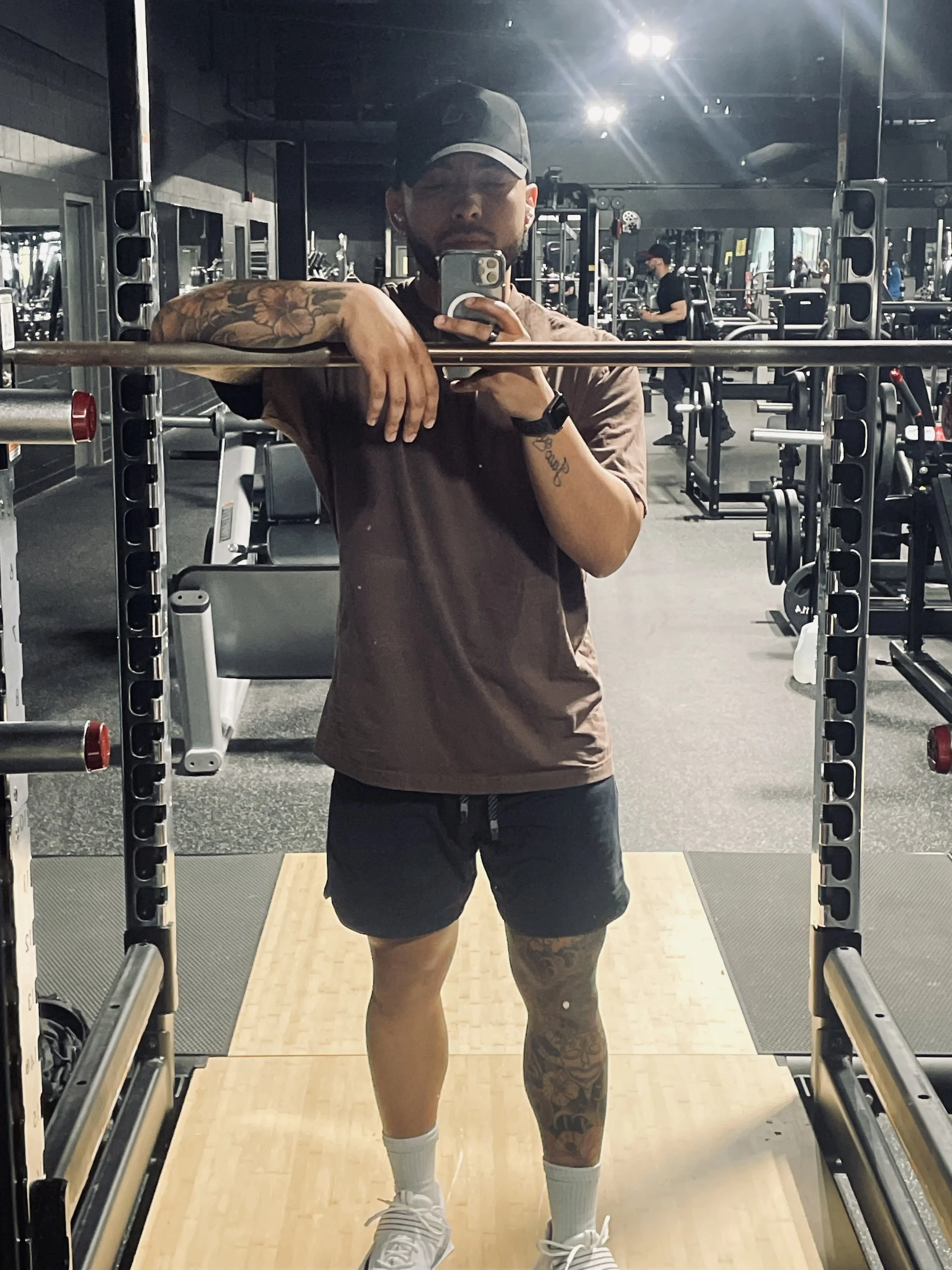 A man taking a selfie in a gym mirror, wearing a brown t-shirt, black shorts, white socks, and white sneakers. He has tattoos on his left arm and right leg, and is wearing a dark cap. The gym is filled with workout equipment and other people exercising in the background.