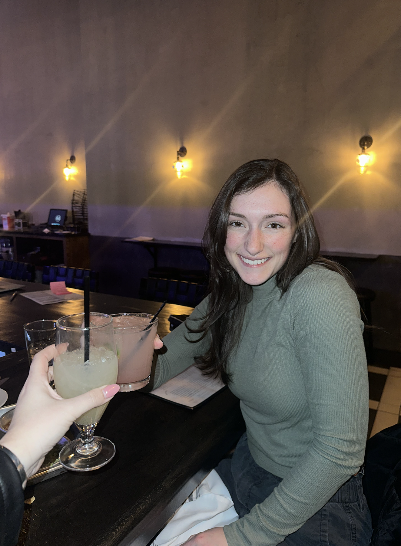 A young woman with long brown hair smiling and sitting at a restaurant table, holding a drink in her hand. There are two drinks on the table in front of her, and the background features warm wall lights.