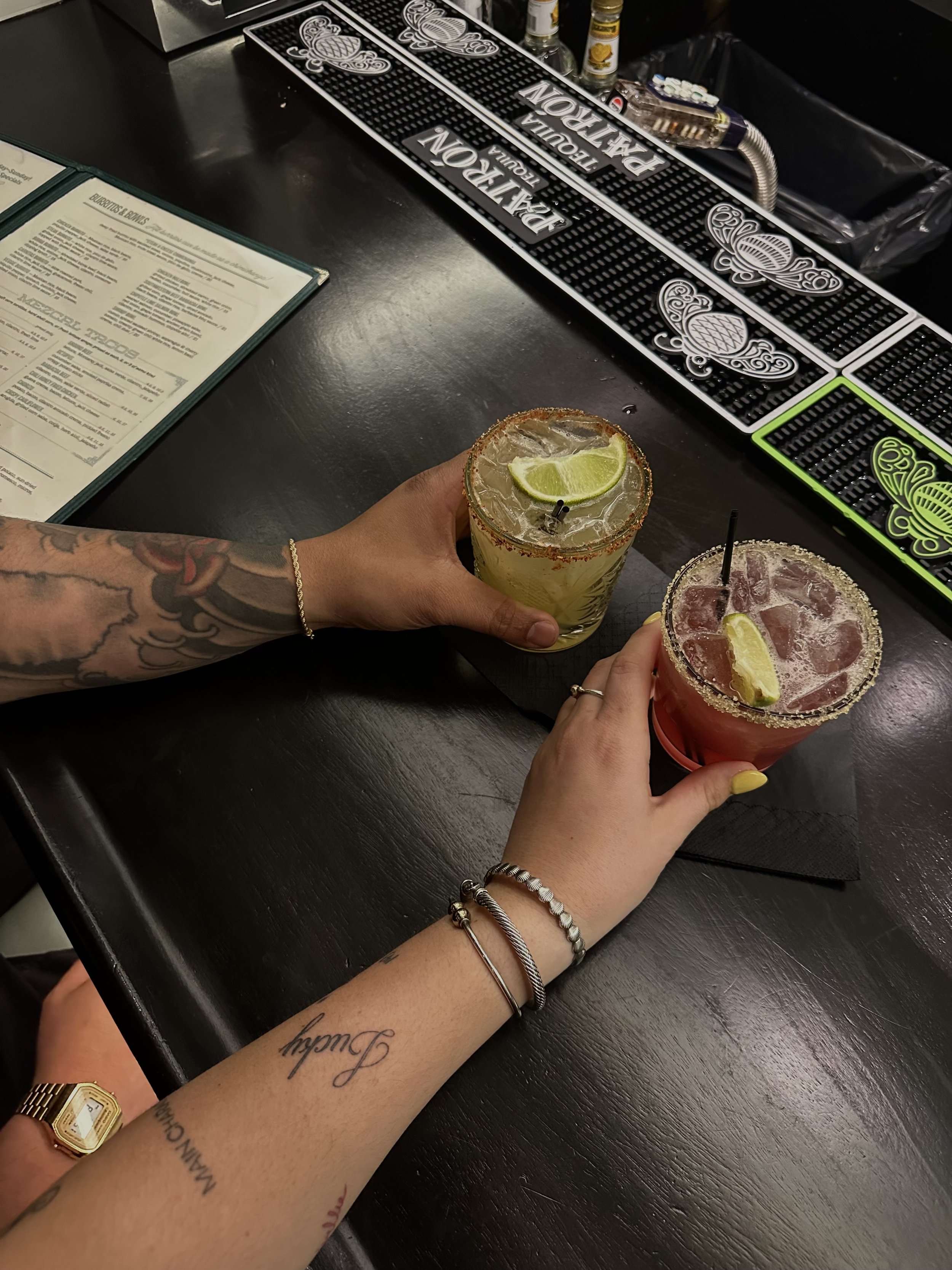 Two hands holding drinks with lime wedges, one with tattoos and jewelry, resting on a dark bar counter. In the background, bar mats and bottles are visible.