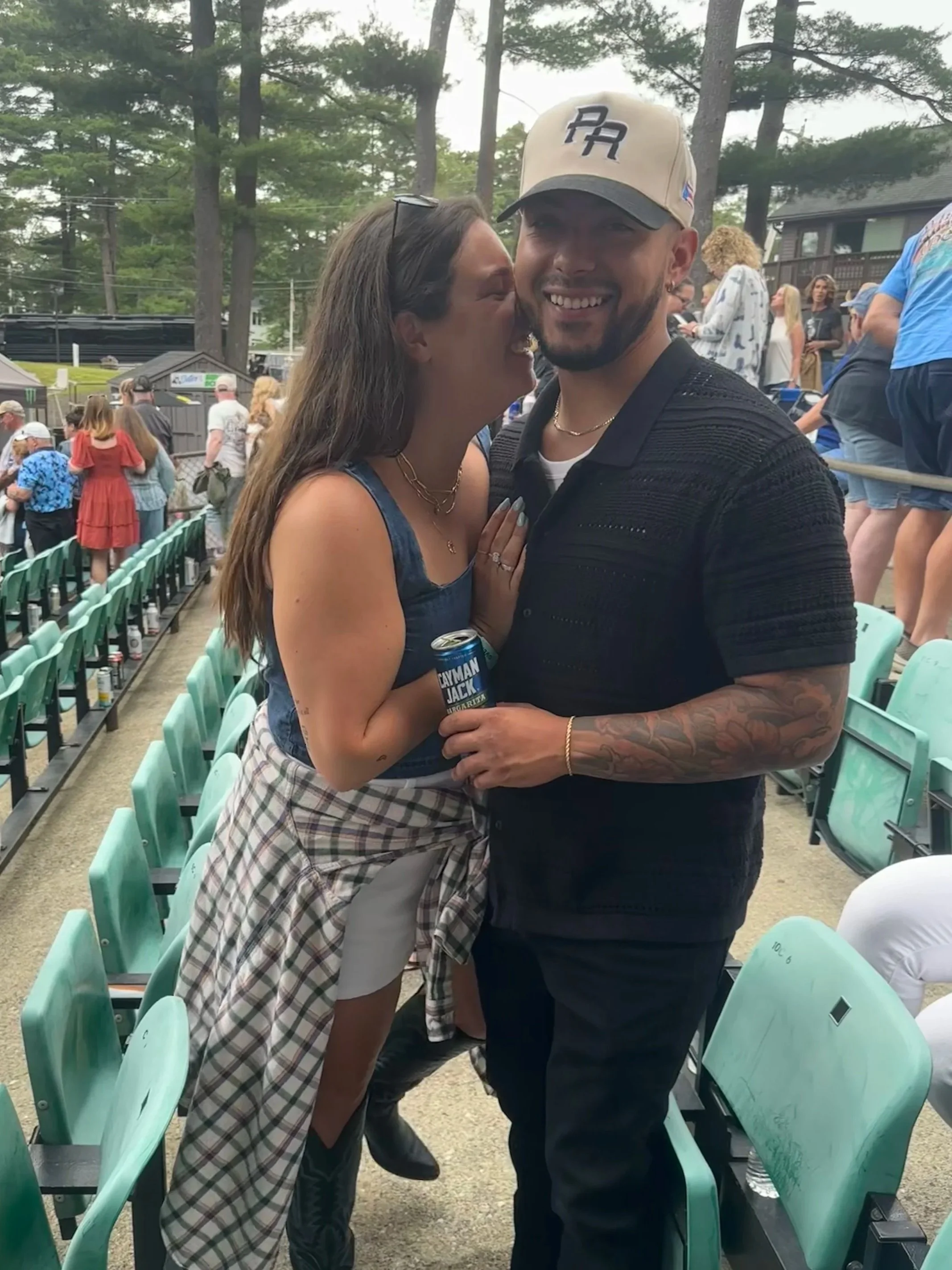 A woman and a man are smiling and hugging at an outdoor event. The woman is wearing a sleeveless top, plaid skirt, and sunglasses on her head, holding a can of Rayman Jack beer. The man is wearing a beige cap, black shirt, and has tattoos on his arm.