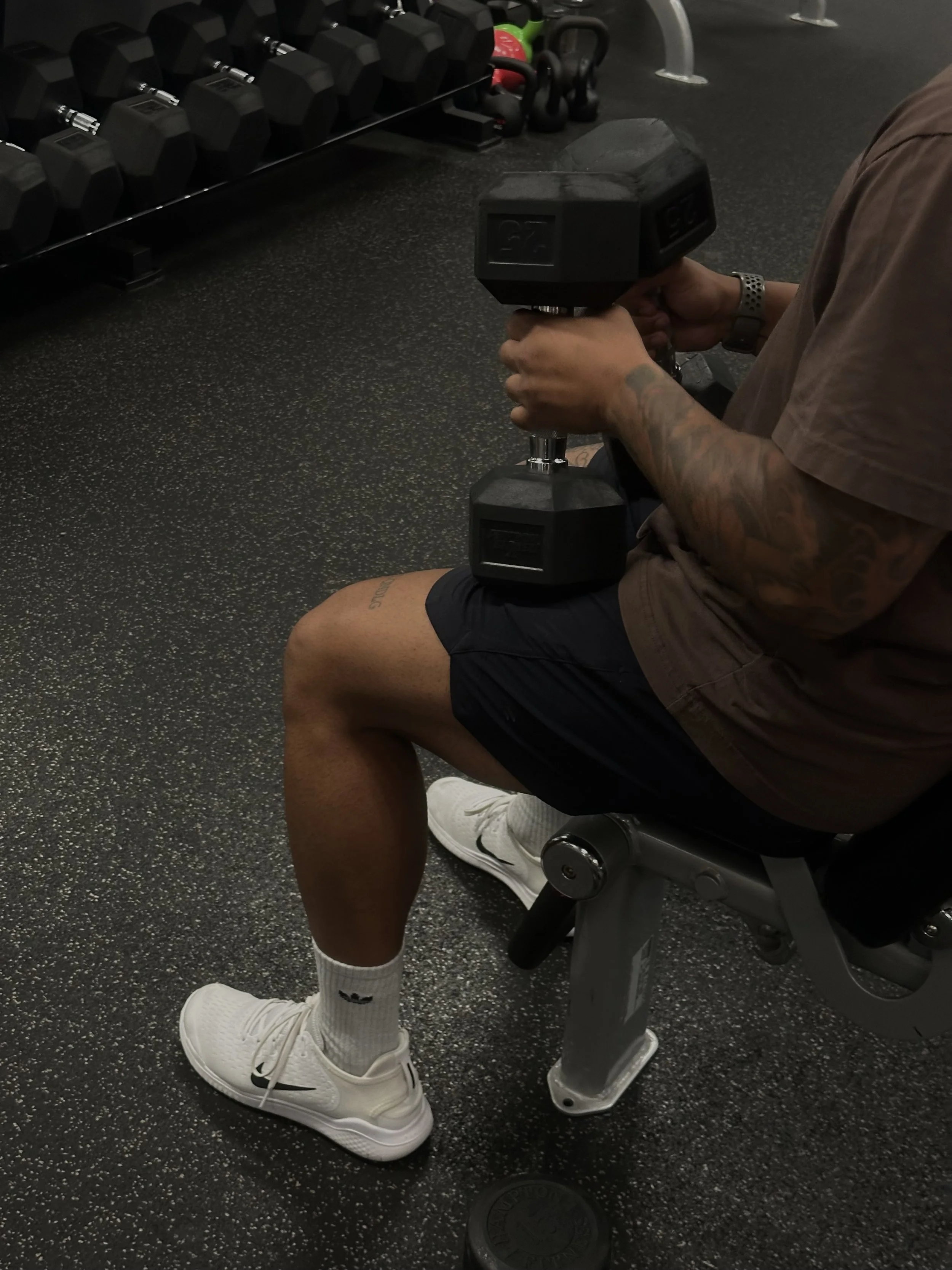 Person sitting on a gym bench holding a 25-pound dumbbell in each hand, wearing a brown t-shirt, black shorts, white Adidas sneakers, and white Nike socks, with a rack of dumbbells and kettlebells visible in the background.