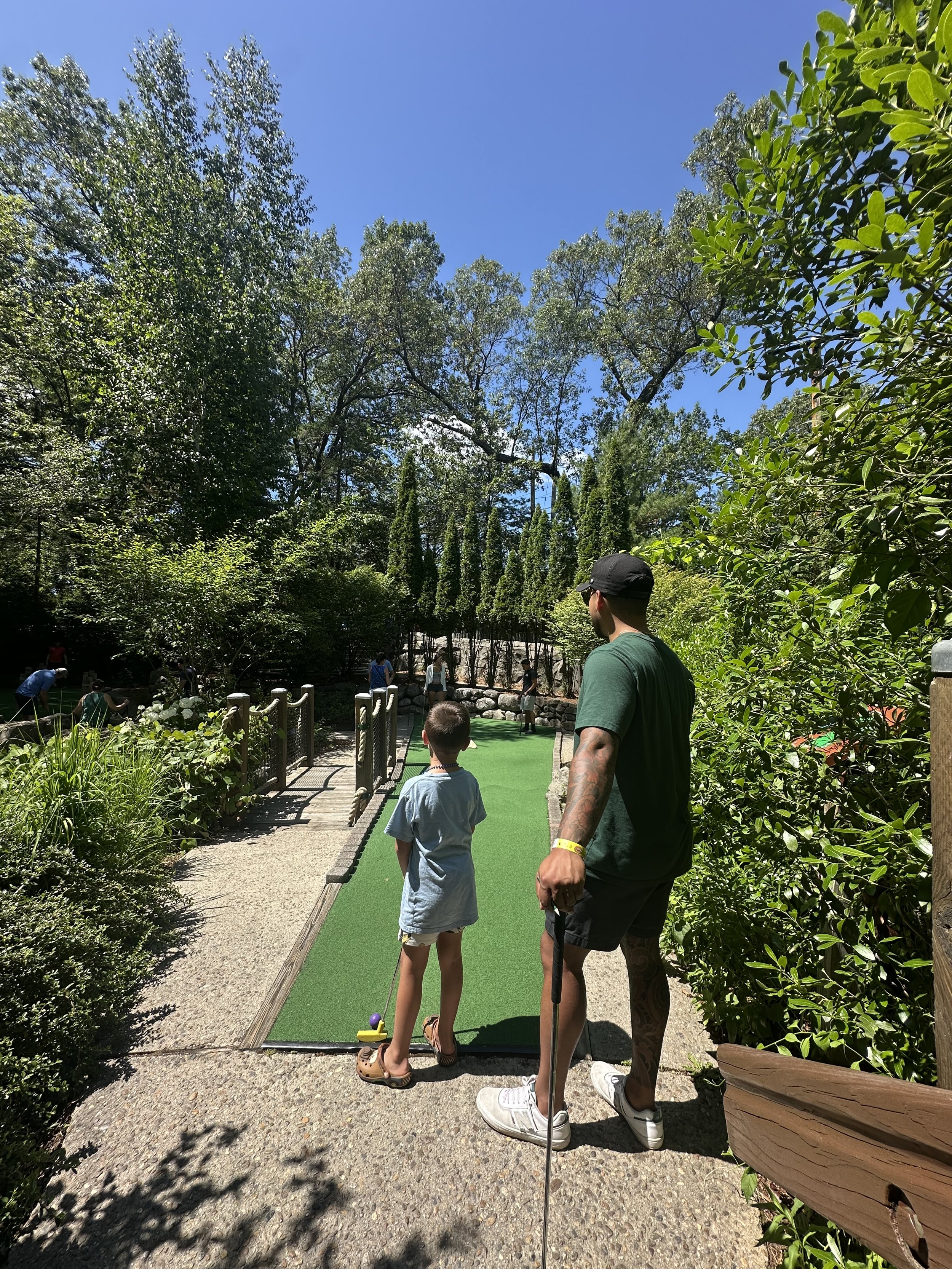 A man and a young girl stand on a mini golf course surrounded by trees and greenery, preparing to take a shot. Other people are visible in the background enjoying the outdoor setting.