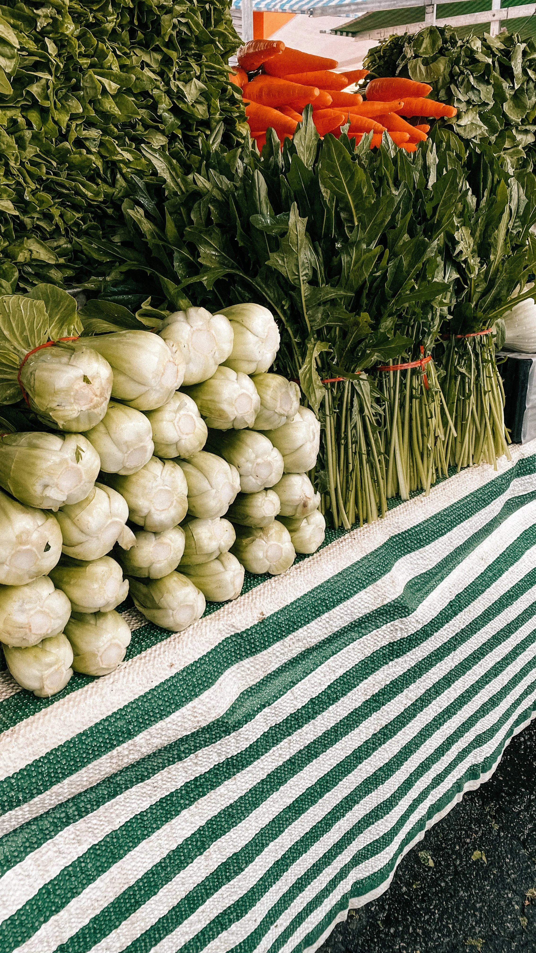 A display of fresh vegetables including white turnips, carrots, and leafy greens on a striped green and white tablecloth at a market.