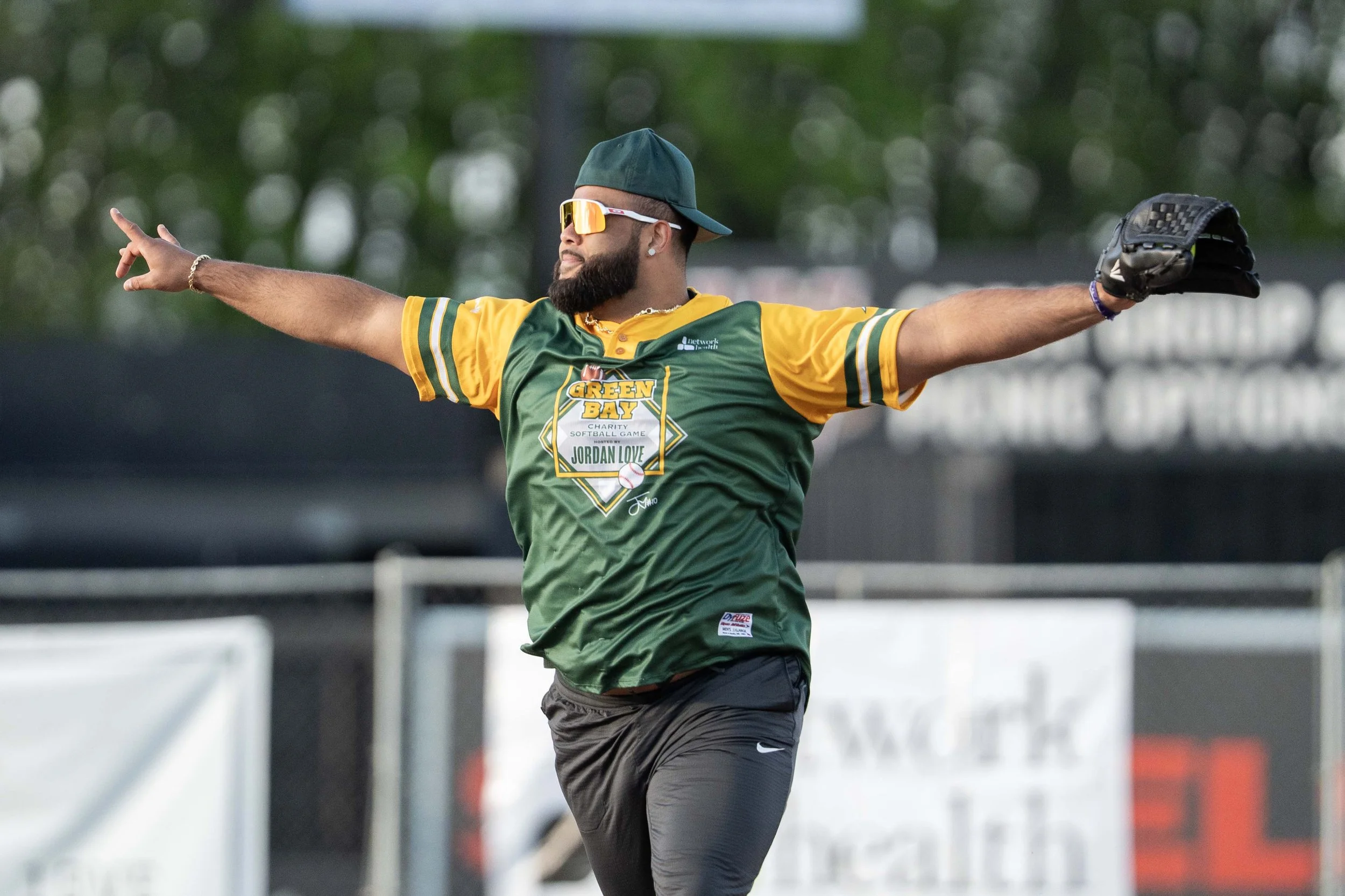 A man wearing a green and yellow Green Bay charity softball game jersey, sunglasses, and a hat is standing on a softball field with arms outstretched and wearing a glove on his left hand.