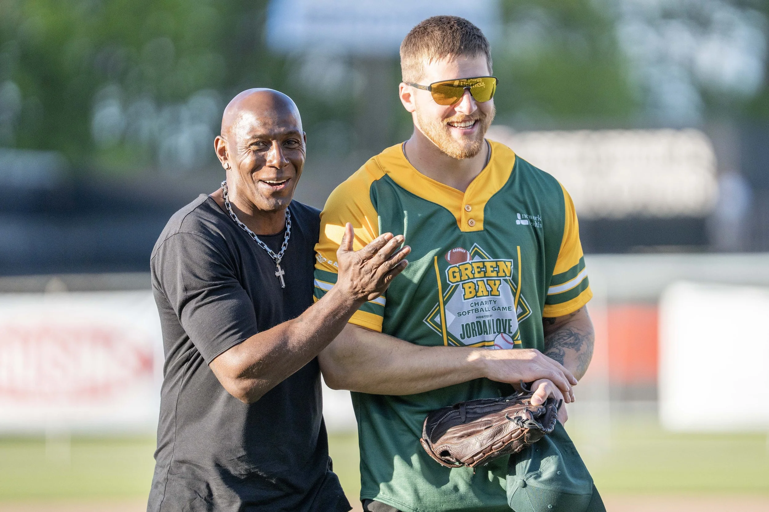 Two smiling baseball players, one with dark skin and a necklace, the other with light skin wearing sunglasses, are standing on a field during daytime.