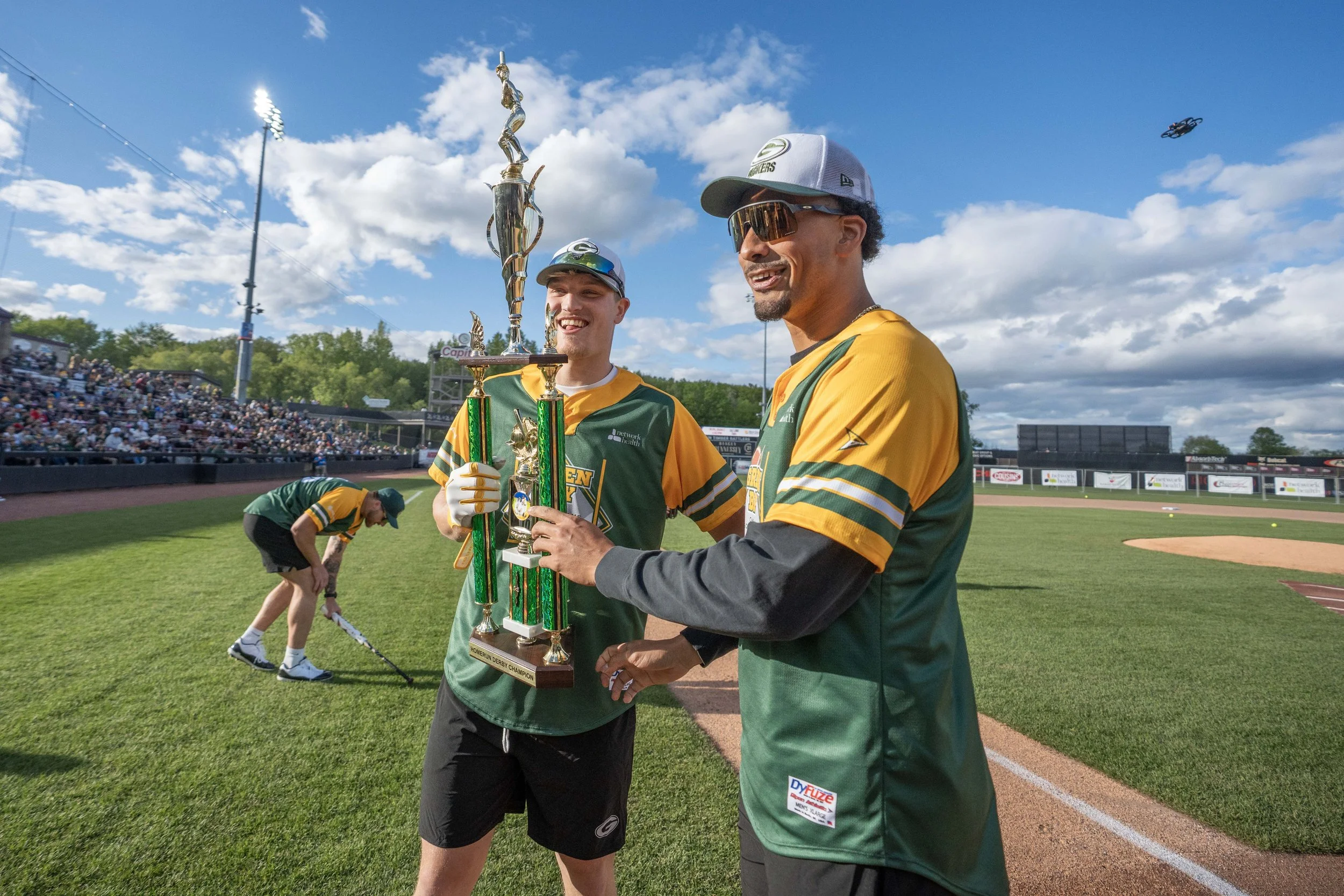 Two baseball players in green and yellow uniforms celebrating with a large trophy on the field, with a crowd in the stands and a person sweeping in the background, under a partly cloudy sky.