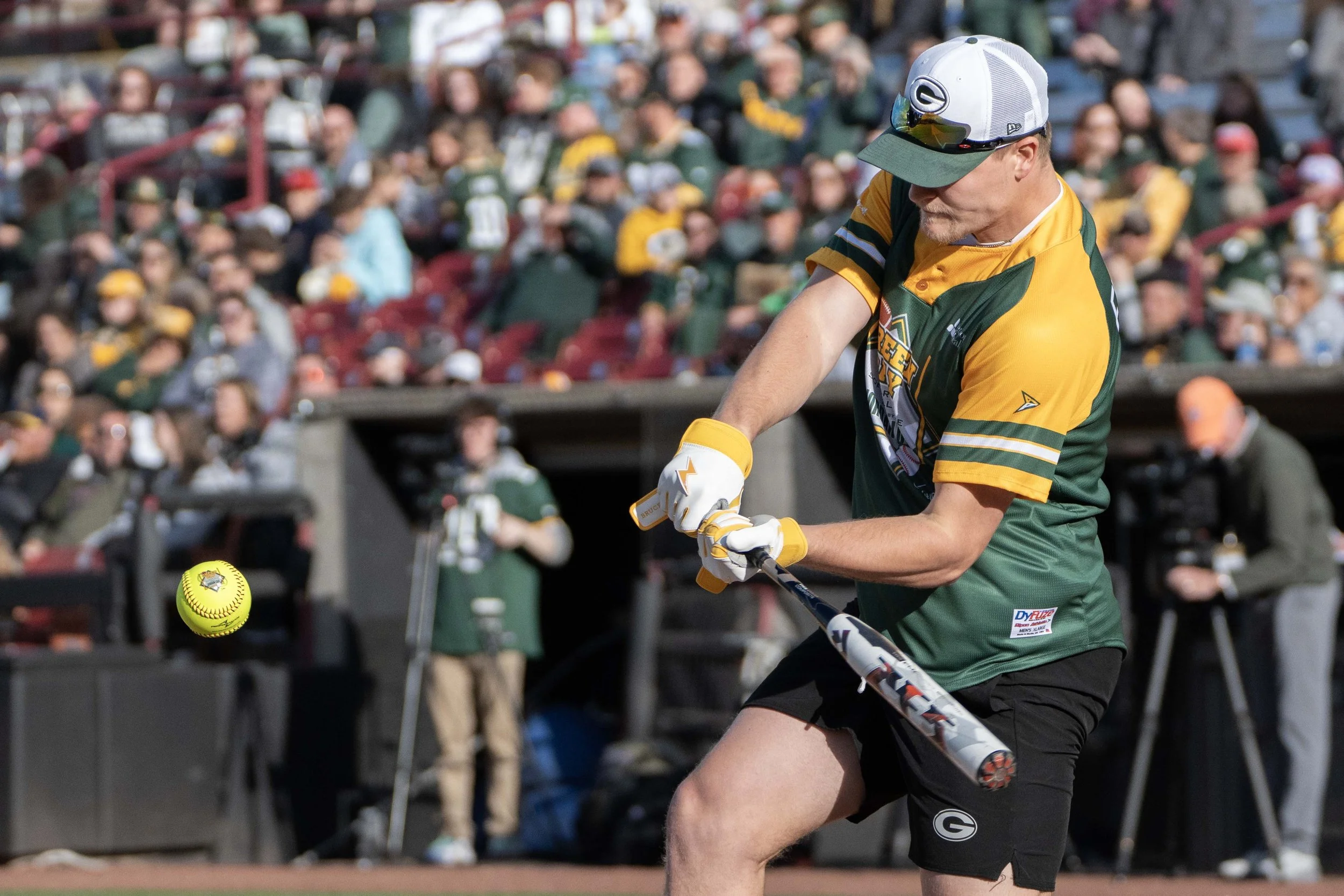 A man in a Green Bay Packers jersey and hat playing softball or baseball outdoors with a crowd in the background.