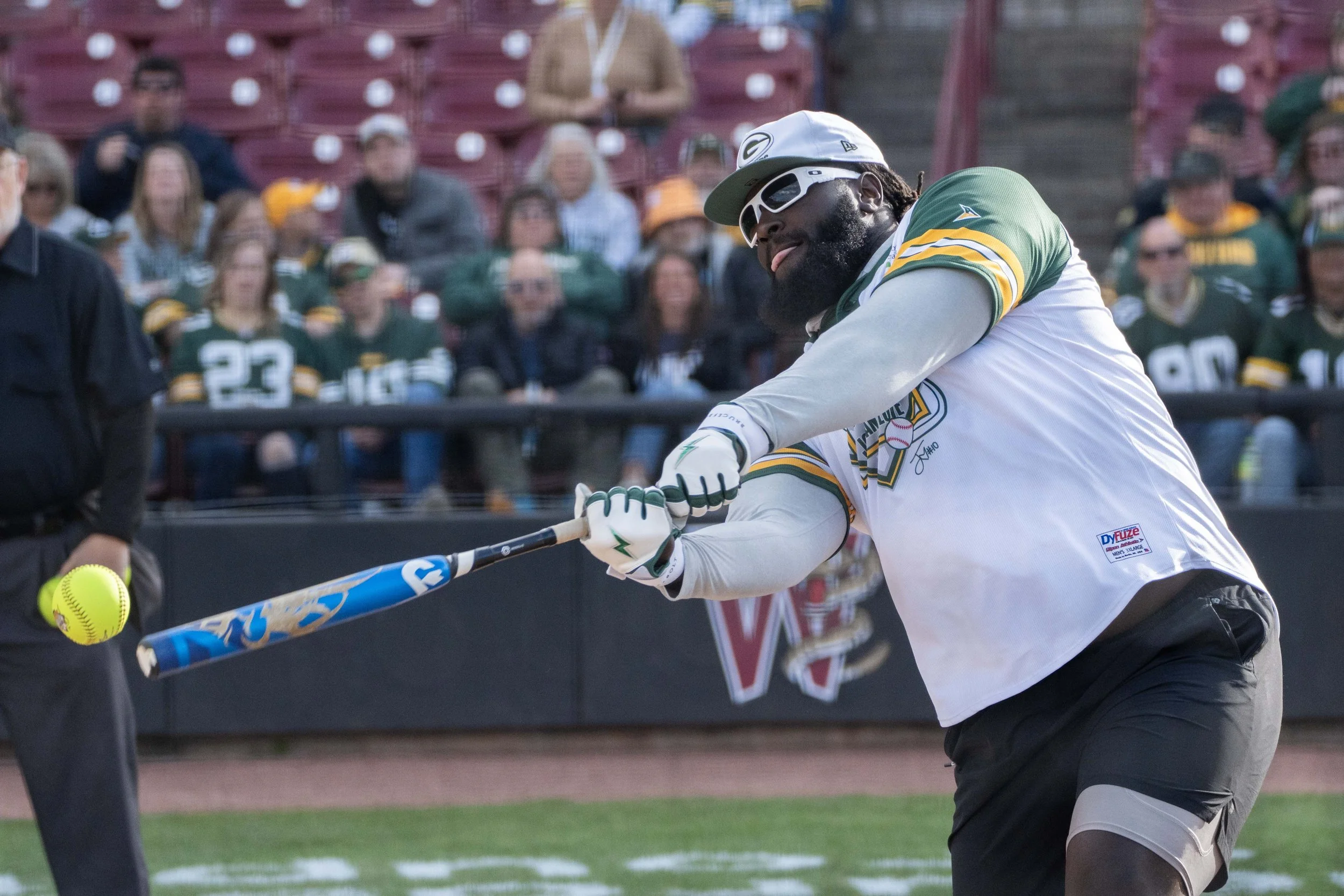 A baseball player in a Green Bay Packers jersey and white gloves hits a yellow softball with a blue bat during a game.