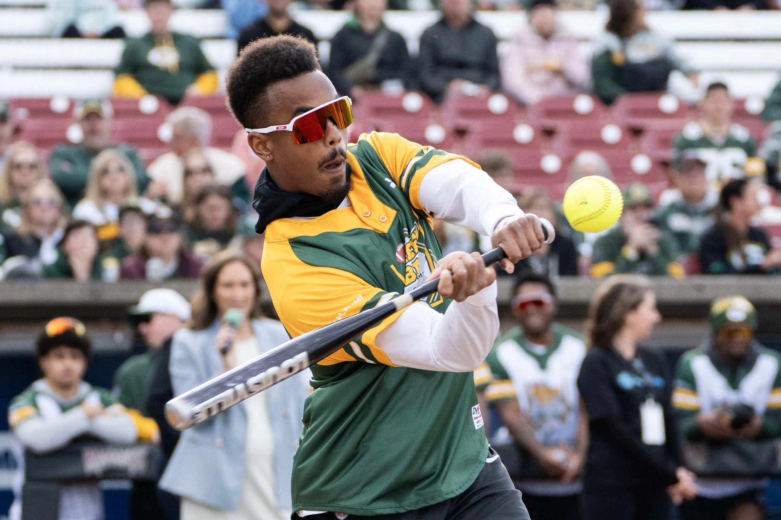 A man wearing sunglasses and a green and yellow sports jersey is swinging a softball bat at a yellow softball during a game. Spectators are watching in the background.