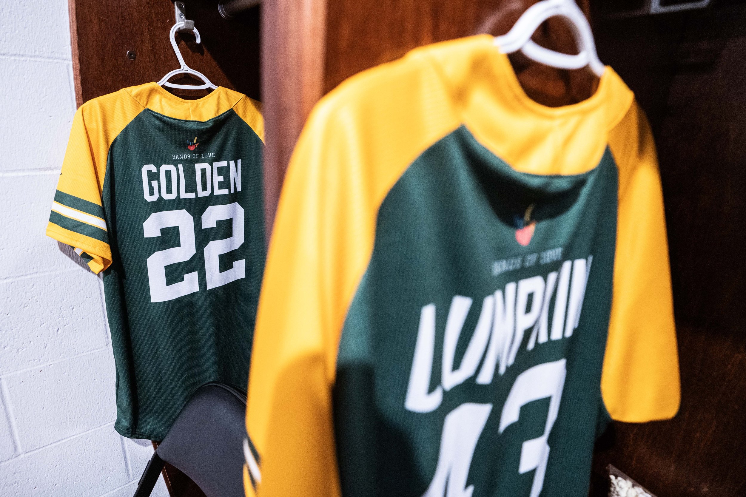 Two green and yellow sports jerseys hanging in a locker room. One has the name 'GOLDEN' and the number '22'. The other shows the word 'LAUNPLIN' and the number '13'.