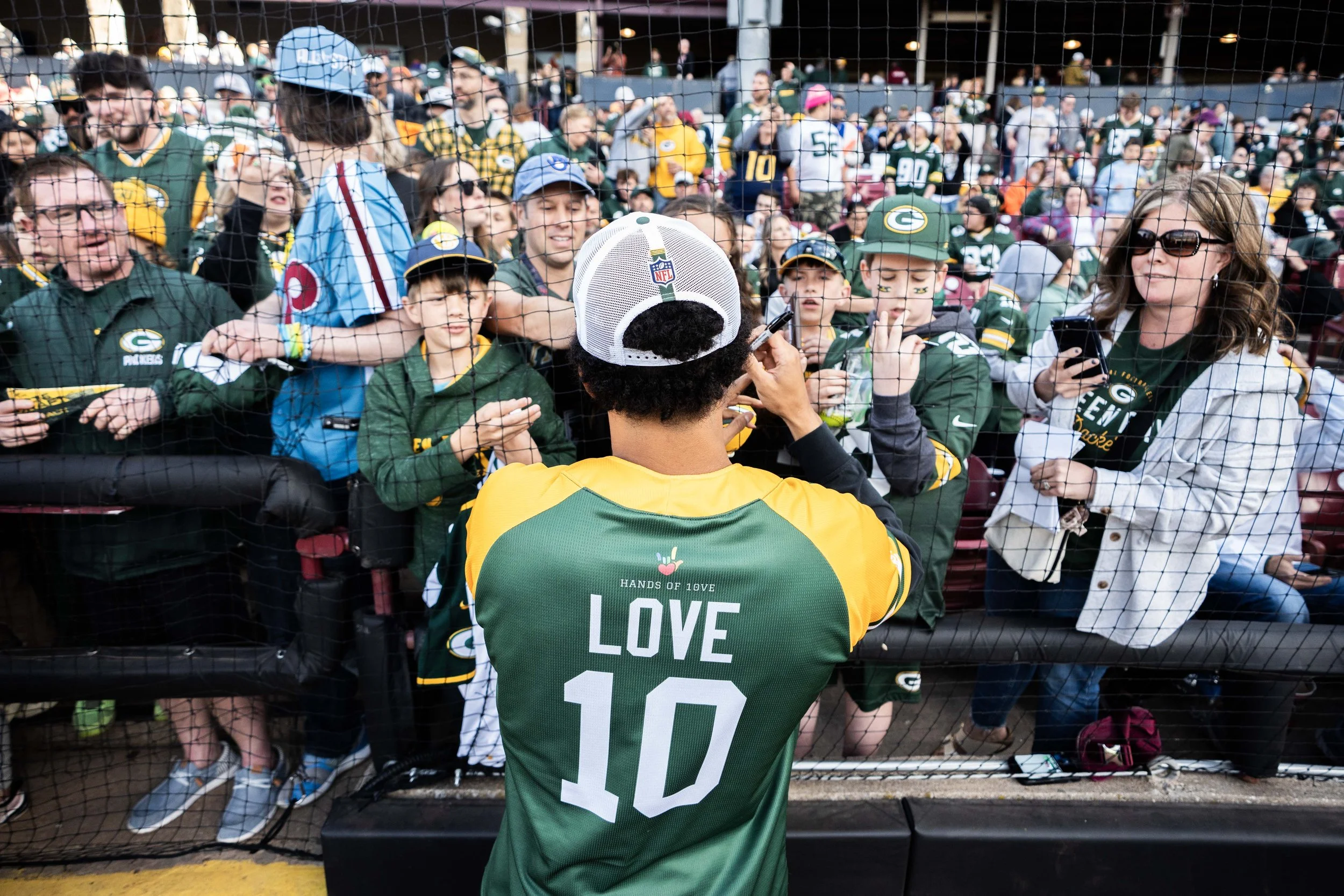 A football player in a green and yellow jersey with the name LOVE and number 10 on the back is signing autographs for fans behind a barrier at a stadium. Fans, mostly children, are wearing Green Bay Packers jerseys and hats, with some holding out foo