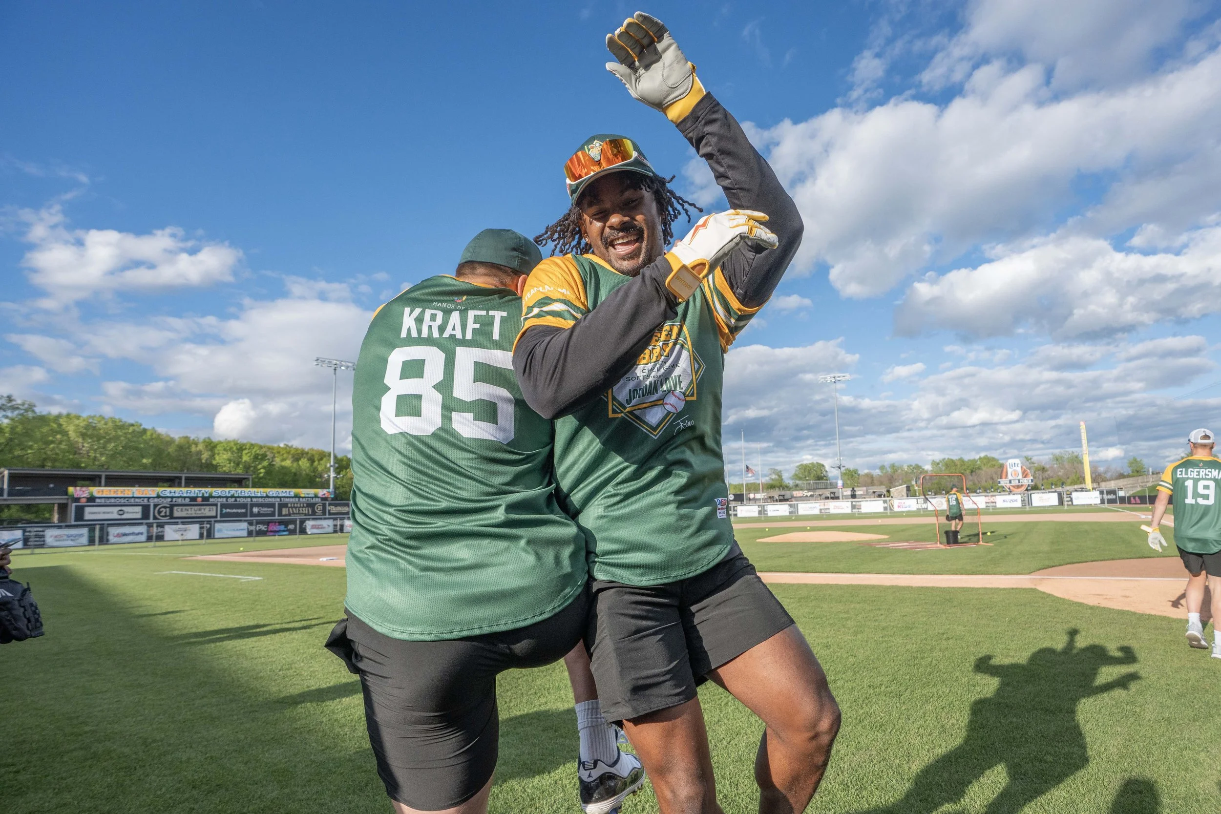Two baseball players celebrating on the field, one with his back to the camera and the other facing forward, both in green jerseys. The one facing forward is smiling and raising his right arm in celebration.