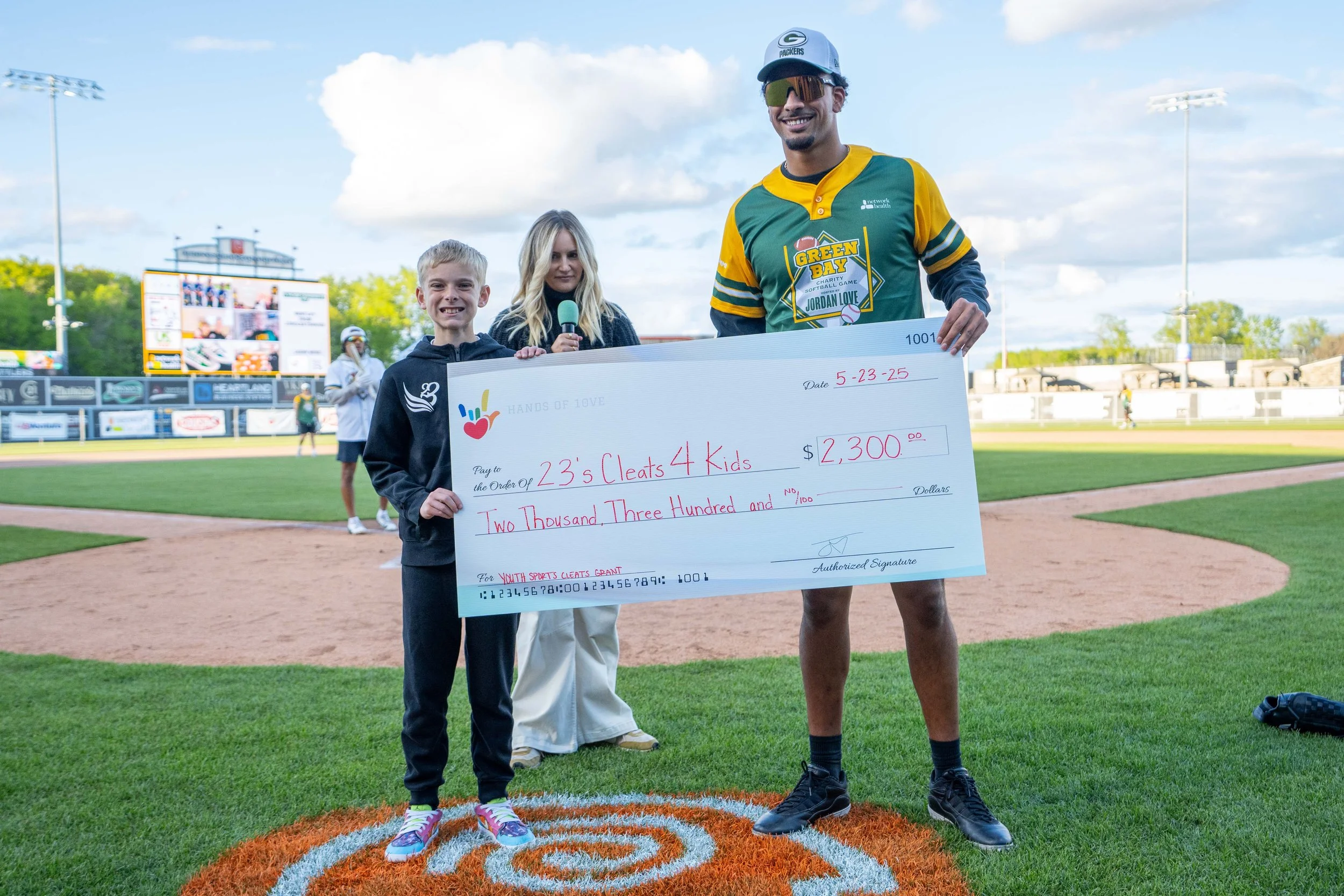 A young boy and an athlete holding a large ceremonial check at a baseball field during a charity event. The boy is smiling, and a woman in the background is holding a microphone. The check is for $2,300 and is made out to '23's Cleats 4 Kids.' The se