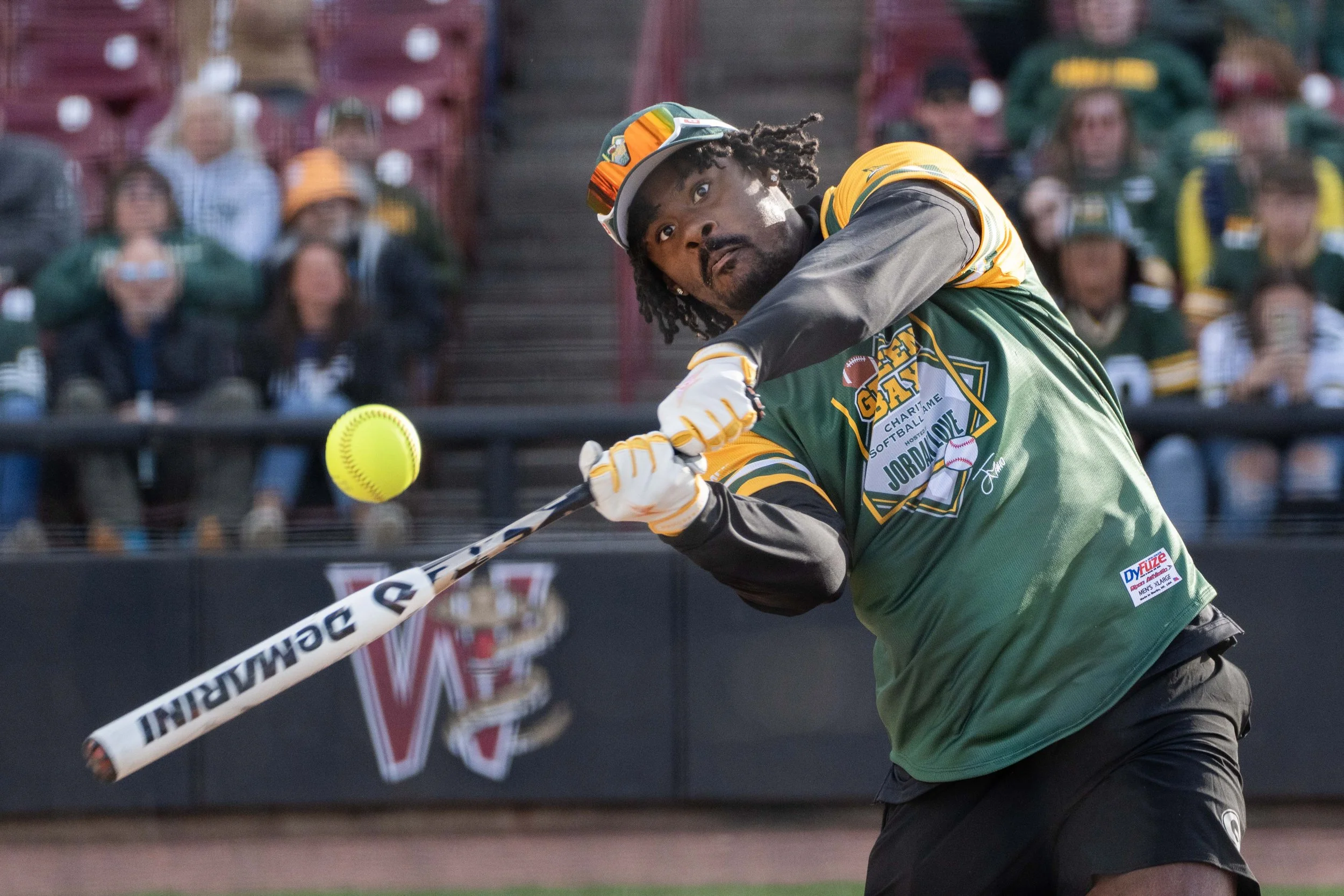 A baseball player in a green and yellow uniform swinging a bat at a yellow softball during a game, with spectators in the stands in the background.